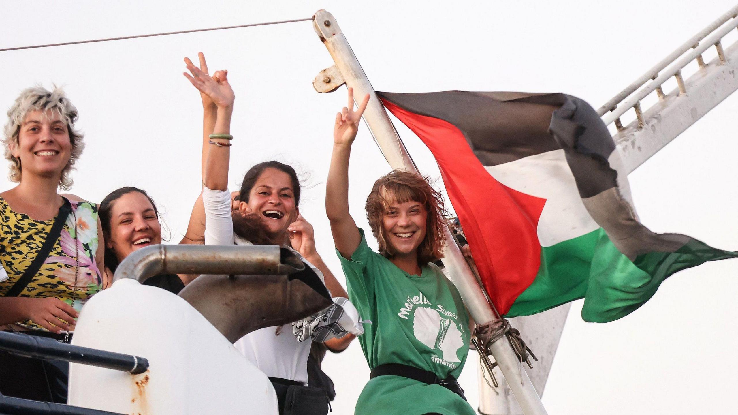 Greta Thunberg and other female crew members of the Global Sumud Flotilla are smiling and waving from a boat.  A Palestinian flag which is tied to the vessel is flying in the breeze.  Greta has short blonde hair and is wearing a green and white t-shirt 