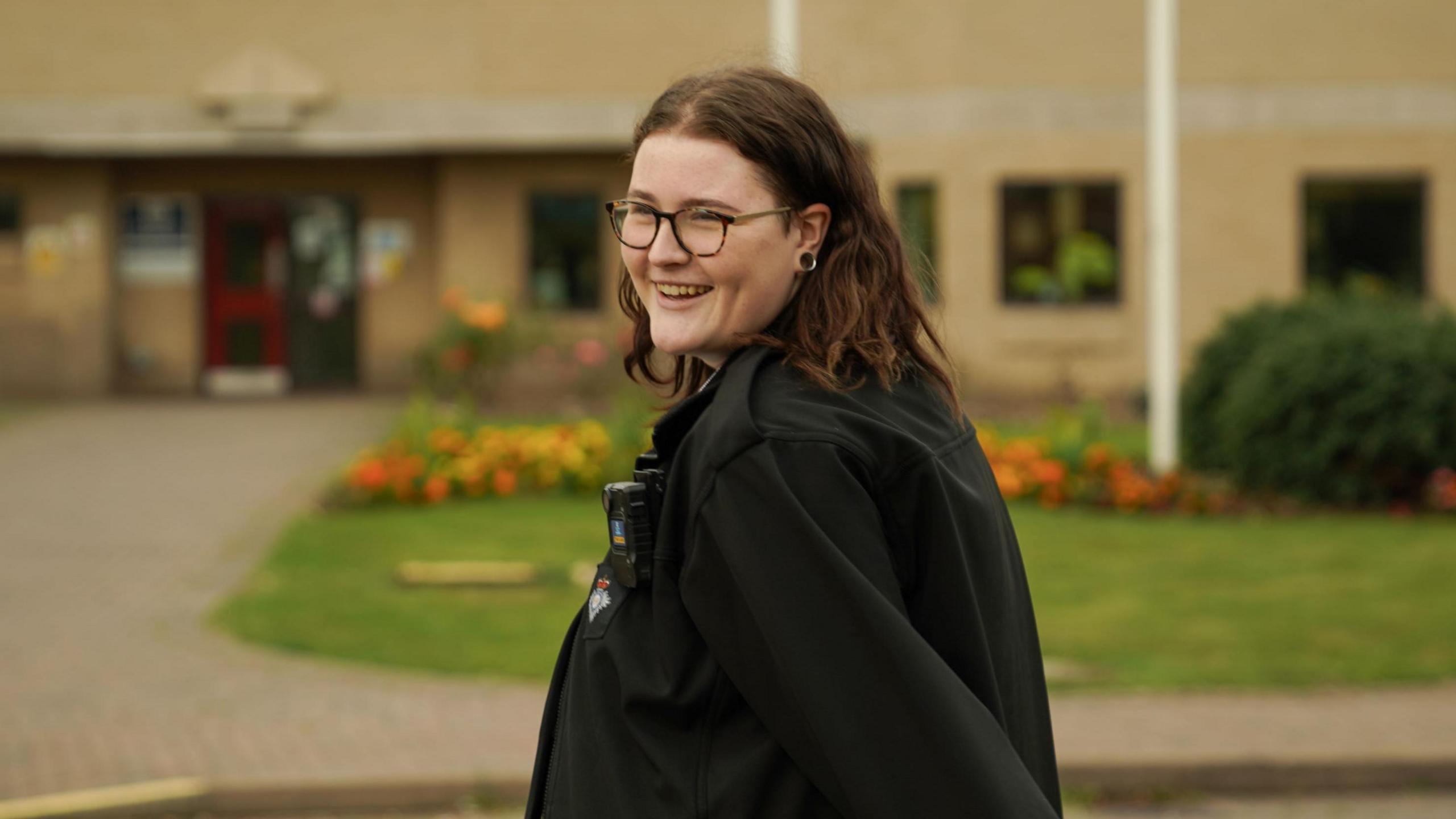 Hollie Peabody pictured outside the prison where they work in their black prison officer uniform. They look over their left shoulder to smile at the camera, surrounded by a neat lawn and a flower bed planted with orange and yellow flowers. 