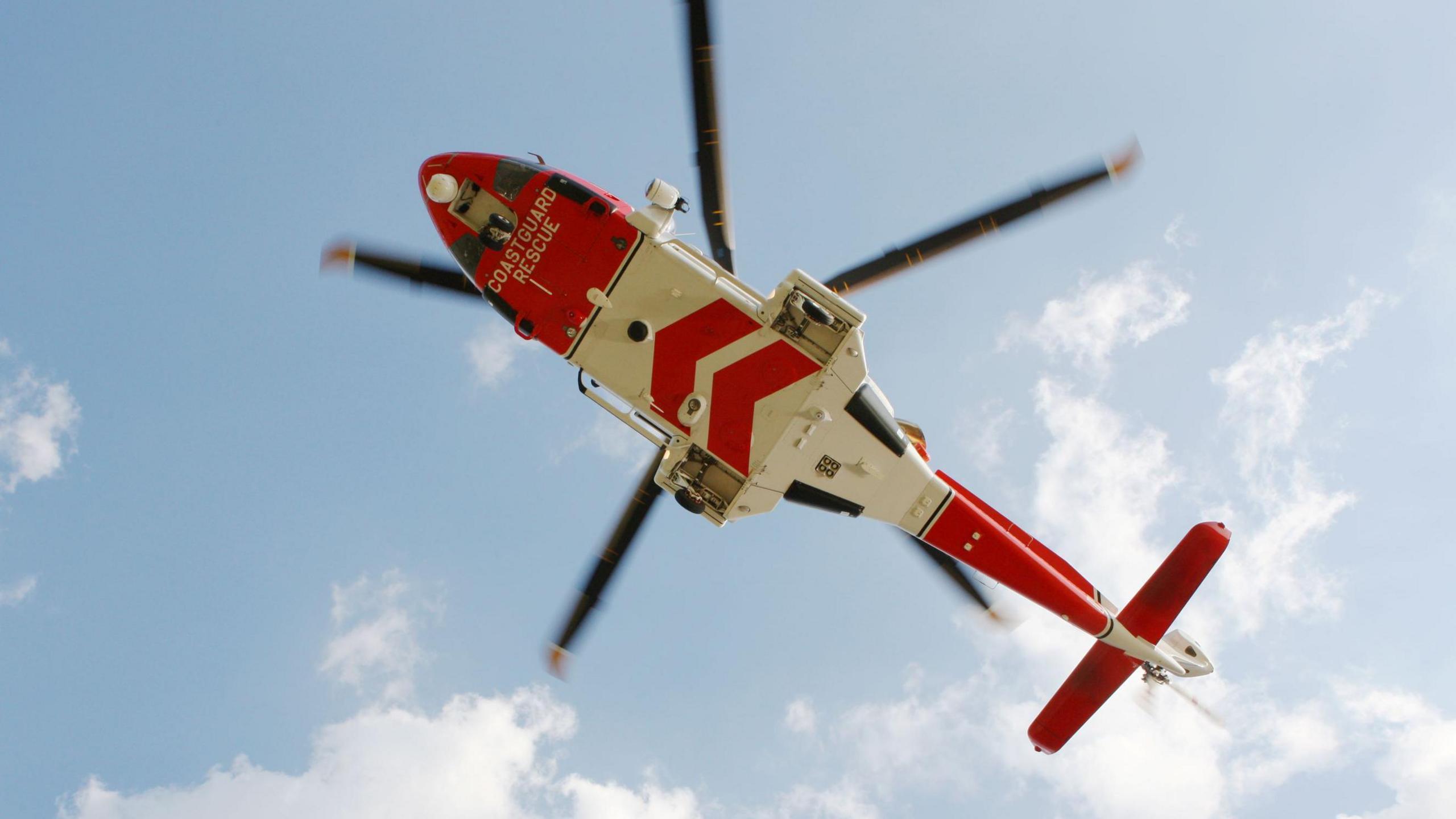 A coastguard helicopter flies overhead. The helicopter is red and white in colour and has its wheels down.