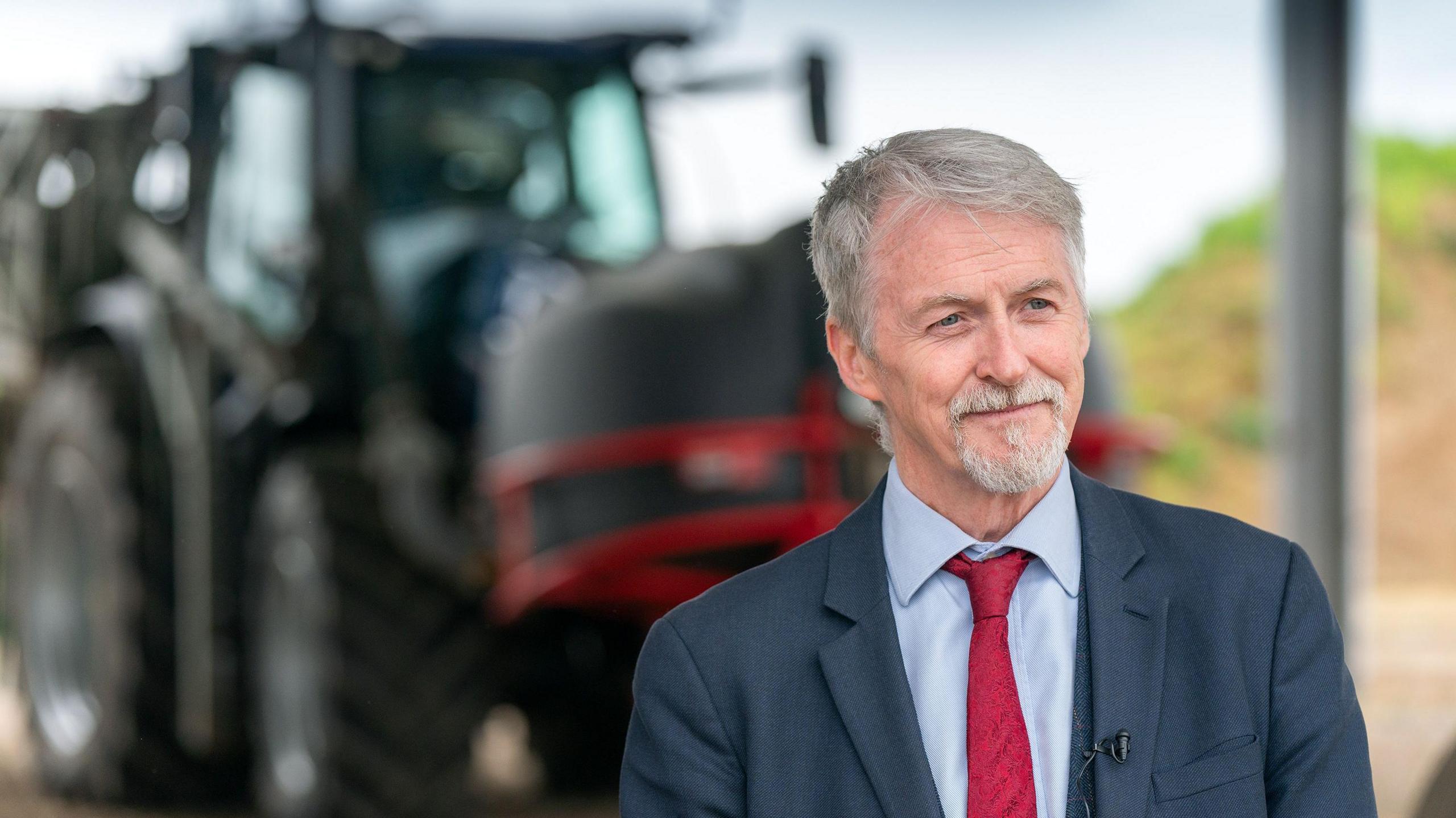 Huw Irranca-Davies, deputy first minister of Wales and cabinet secretary for rural affairs. He has grey hair and a beard and is wearing a blue suit, a blue shirt and red tie. He is standing on a farm, in front of a tractor.