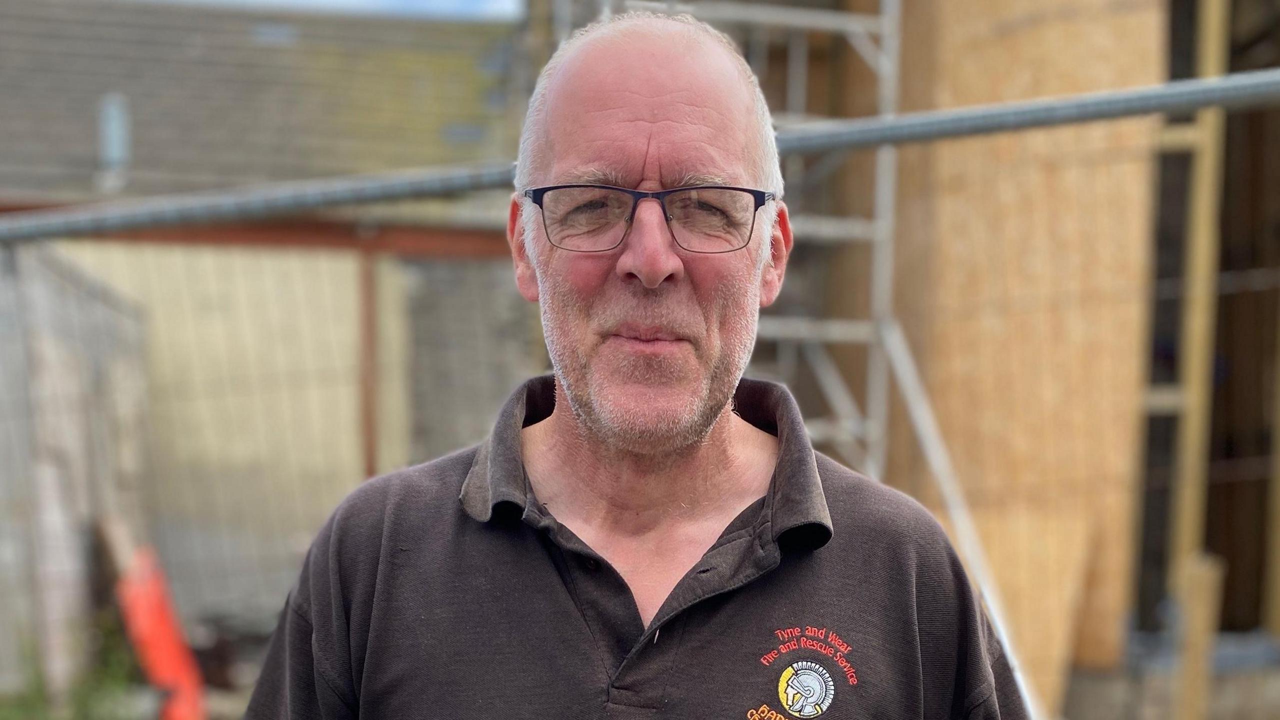 Shaun wears thin-rimmed rectangular glasses and has wispy grey hair and stubble. He's got a brown polo shirt on.