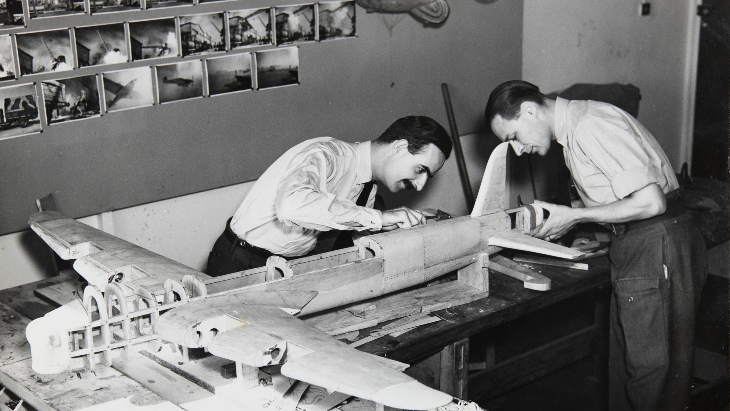 John Howell and Ray Morse are leaning over a model plane in a workshop. They are in trousers and shirts, wearing ties. They appear to be carrying out maintenance or checks of the model.