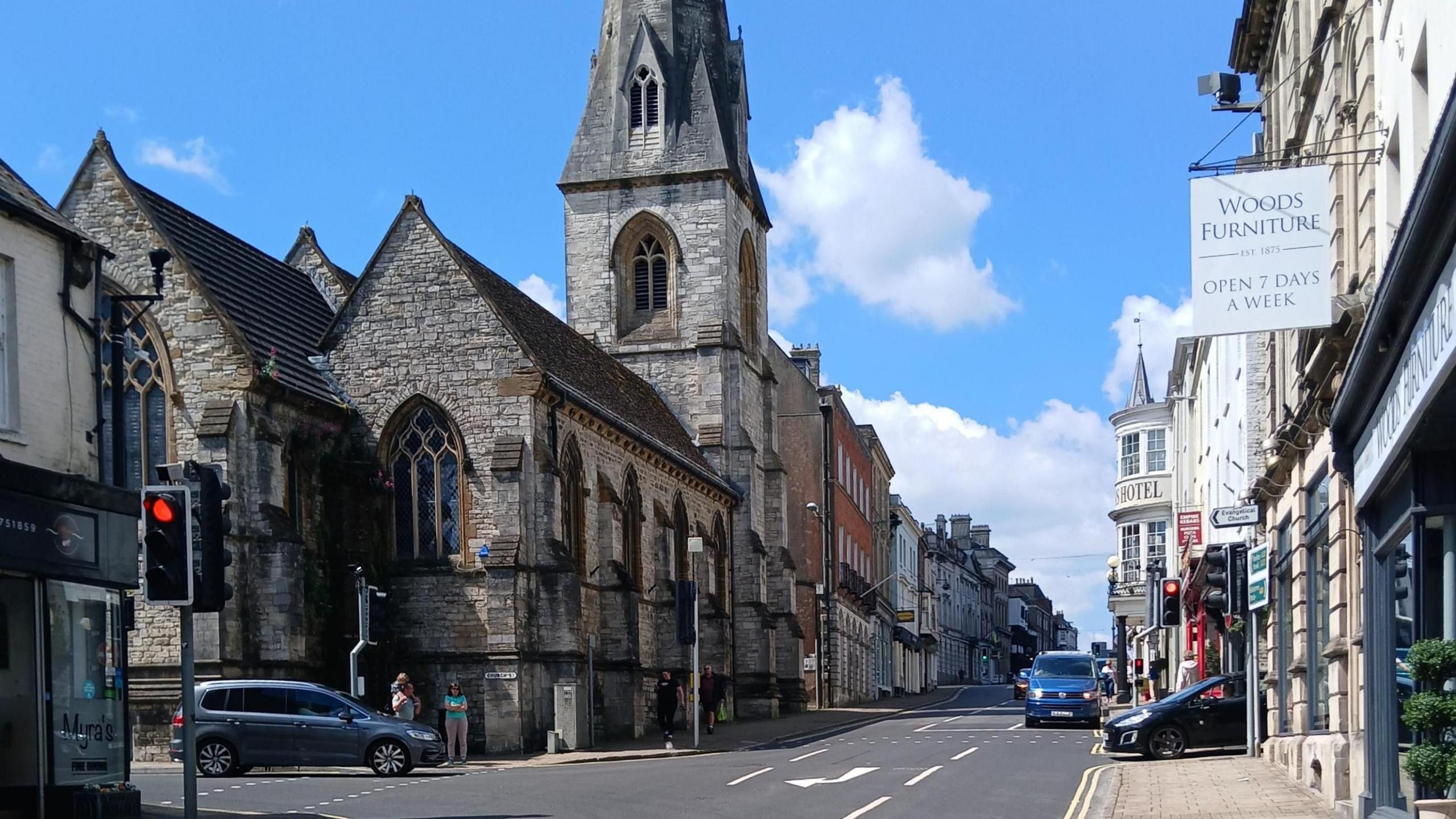 A view of Dorchester High Street, with a grey brick church and church tower, a traffic light to the left and shops along a street to the right. There are cars driving along the road.