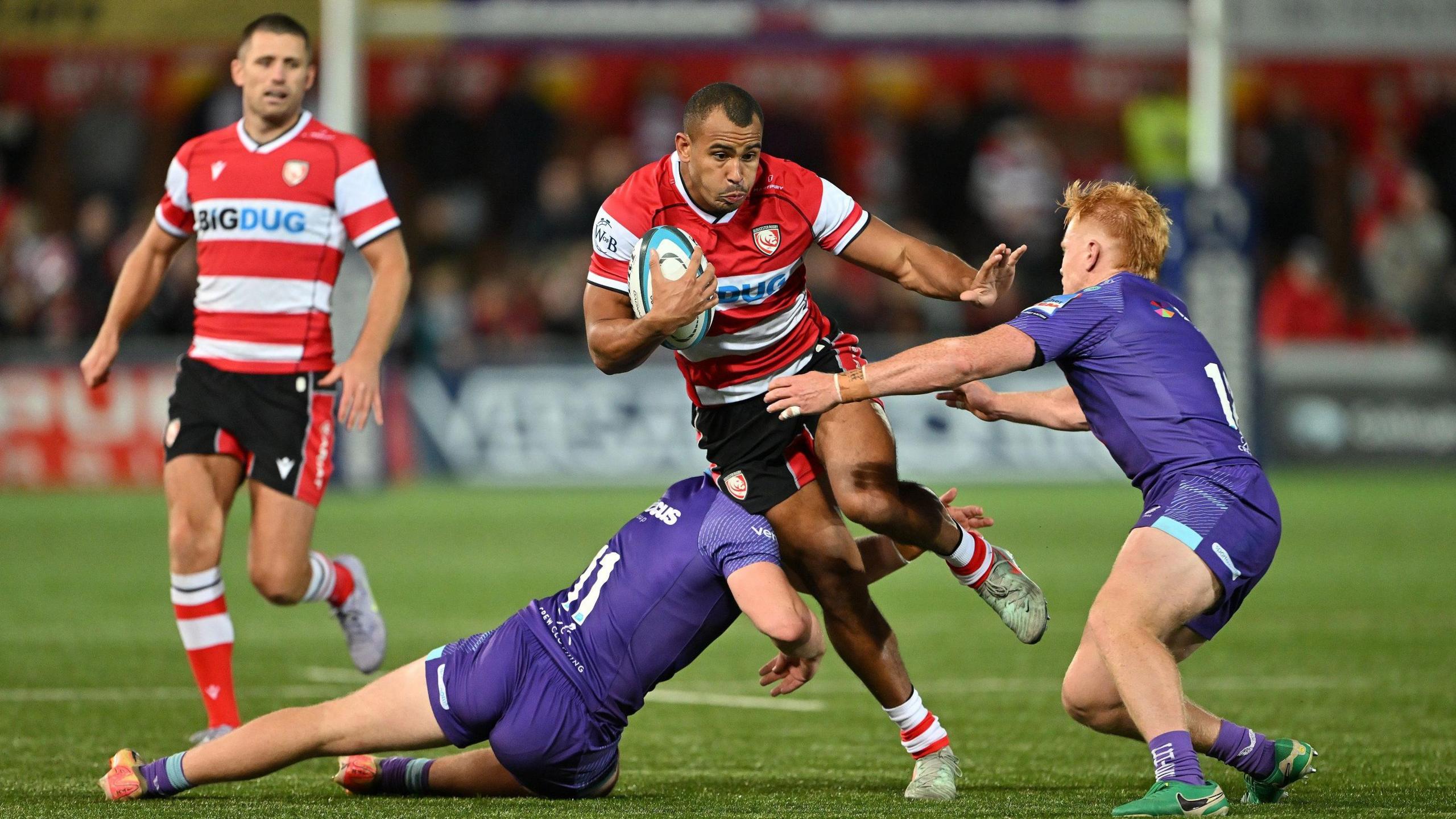A Gloucester Rugby player, wearing a white and red striped top, shields himself from two tackling players from the Exeter Chiefs as he darts between the two, holding his hand up to the face of one, while one of his teammates watches on.