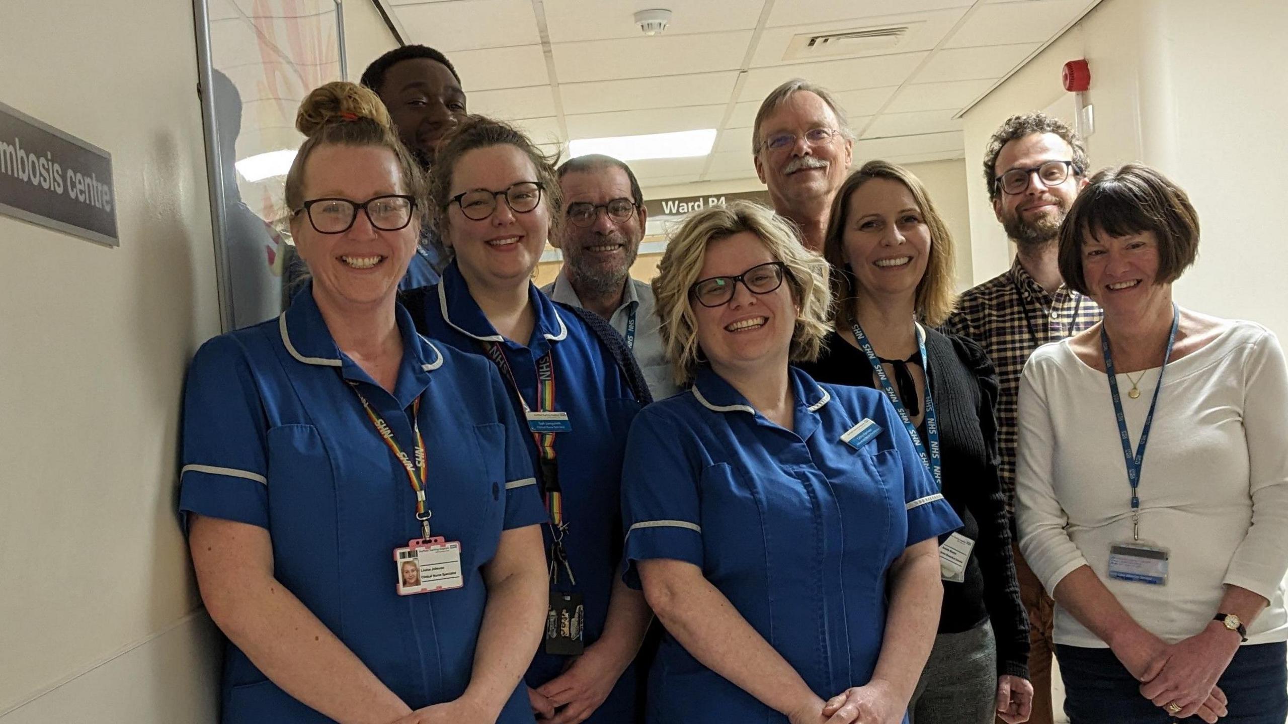 Nine people, some wearing blue scrubs, smile at the camera. They are all standing in a corridor and looking towards the camera. Cathy Harrison is in the middle of the people.