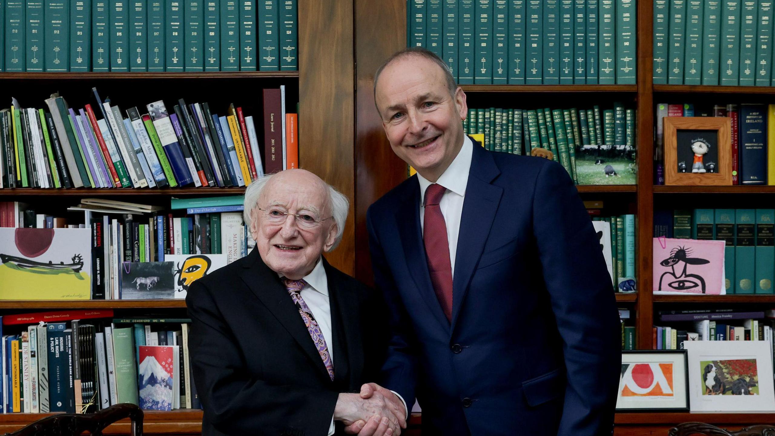 
Michael D Higgins - a shorter man with white hair and glasses who is wearing a dark suit jacket, a patterned tie and a white collared shirt shakes the hand of Micheál Martin, who is wearing a navy suit jacket, a white collared shirt and a red tie. They are standing in a well decorated state room with a Irish tricolour in the background.
