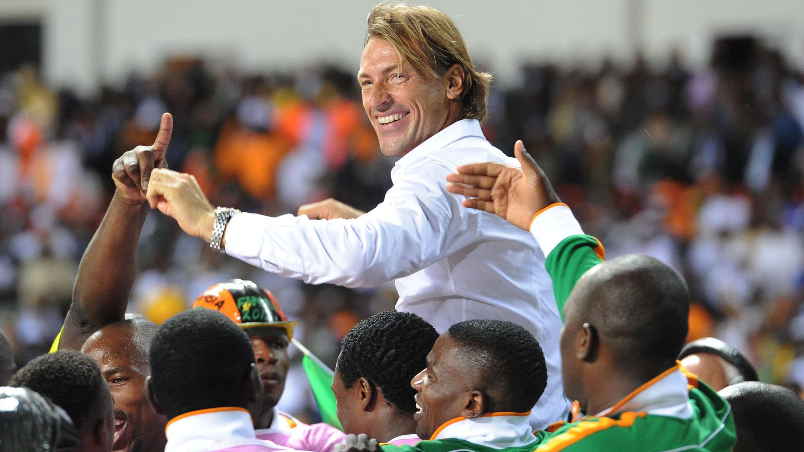 Herve Renard, wearing a white shirt, smiles as he is held aloft by Zambia players celebrating the 2012 Africa Cup of Nations triumph. A packed stand at a football ground is out of focus in the background