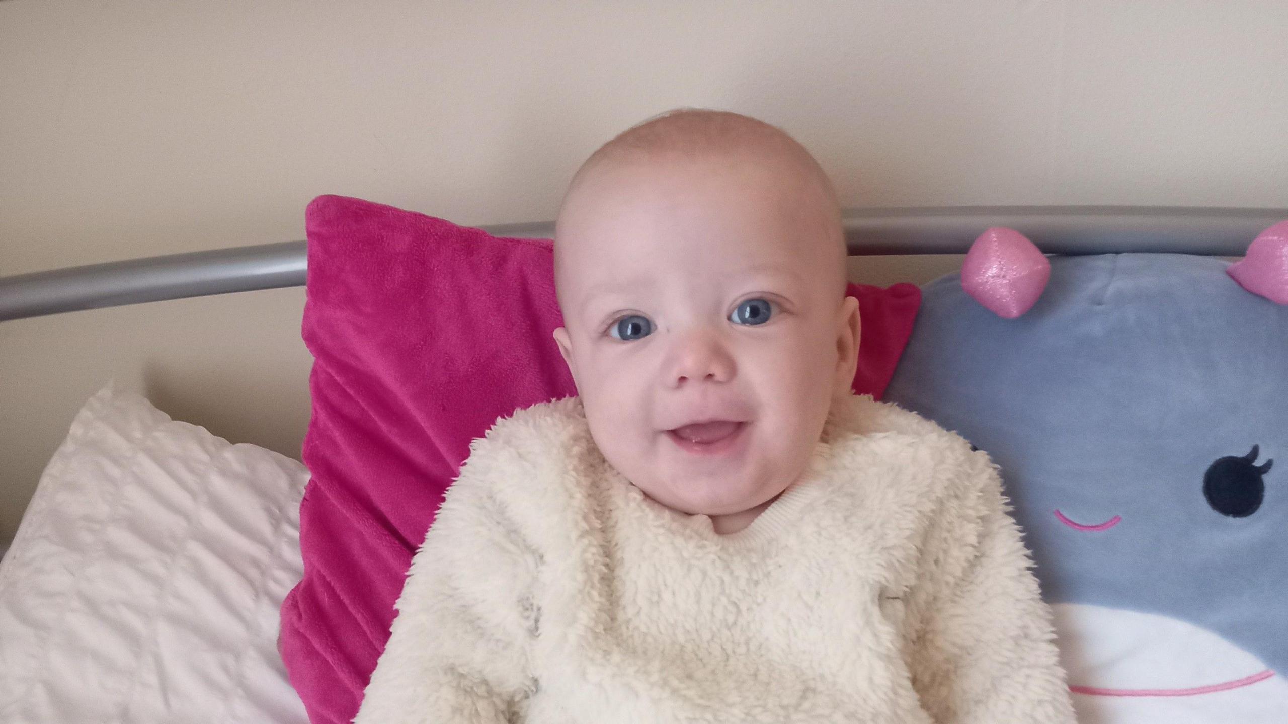 Archie Woodbridge: A blue-eyed, bald baby looking at the camera with his eyes shining and his mouth open. He is wearing a fluffy cream top and is propped up on a pink cushion and a blue-and-white cuddly toy.