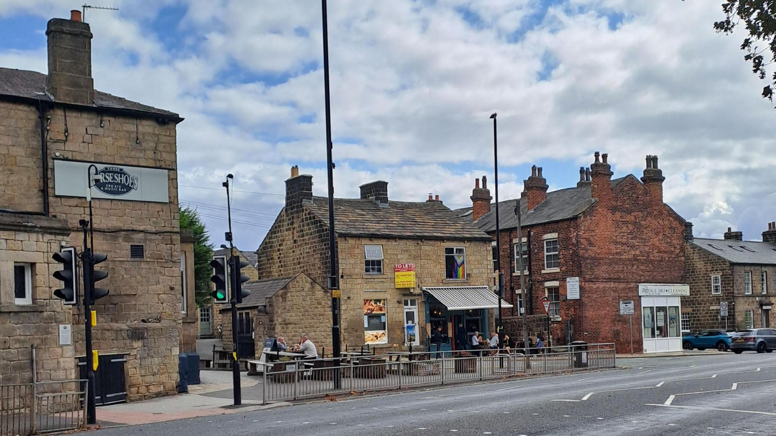 A city street pictured from the other side of a road, featuring bars with punters sat outside. Other street furniture is visible.