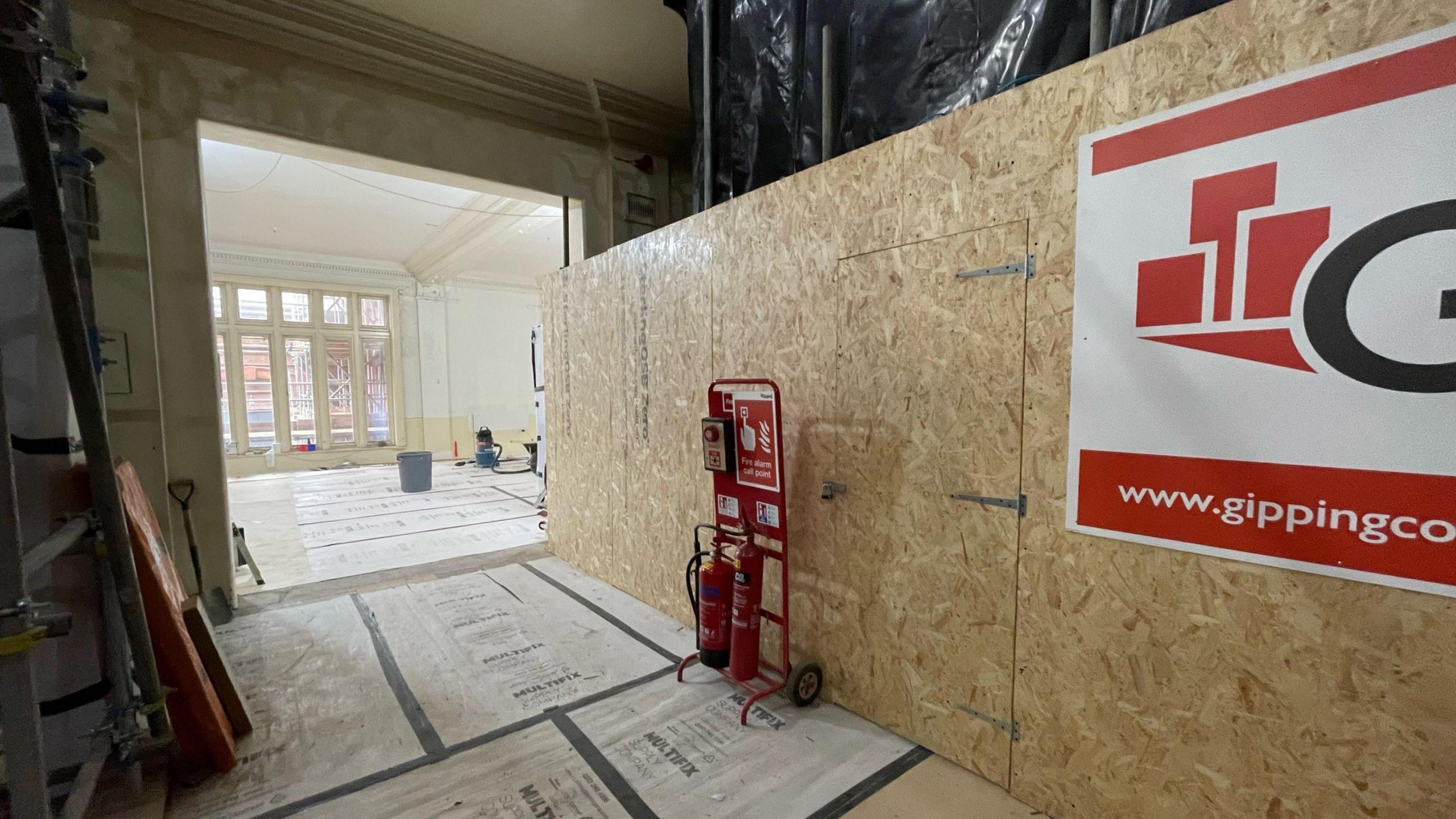 The photo shows a part of the museum and an adjoining room. Construction materials can be seen resting along the walls while a protective surface covers the flood. A bucket can be seen in the background along with several windows.