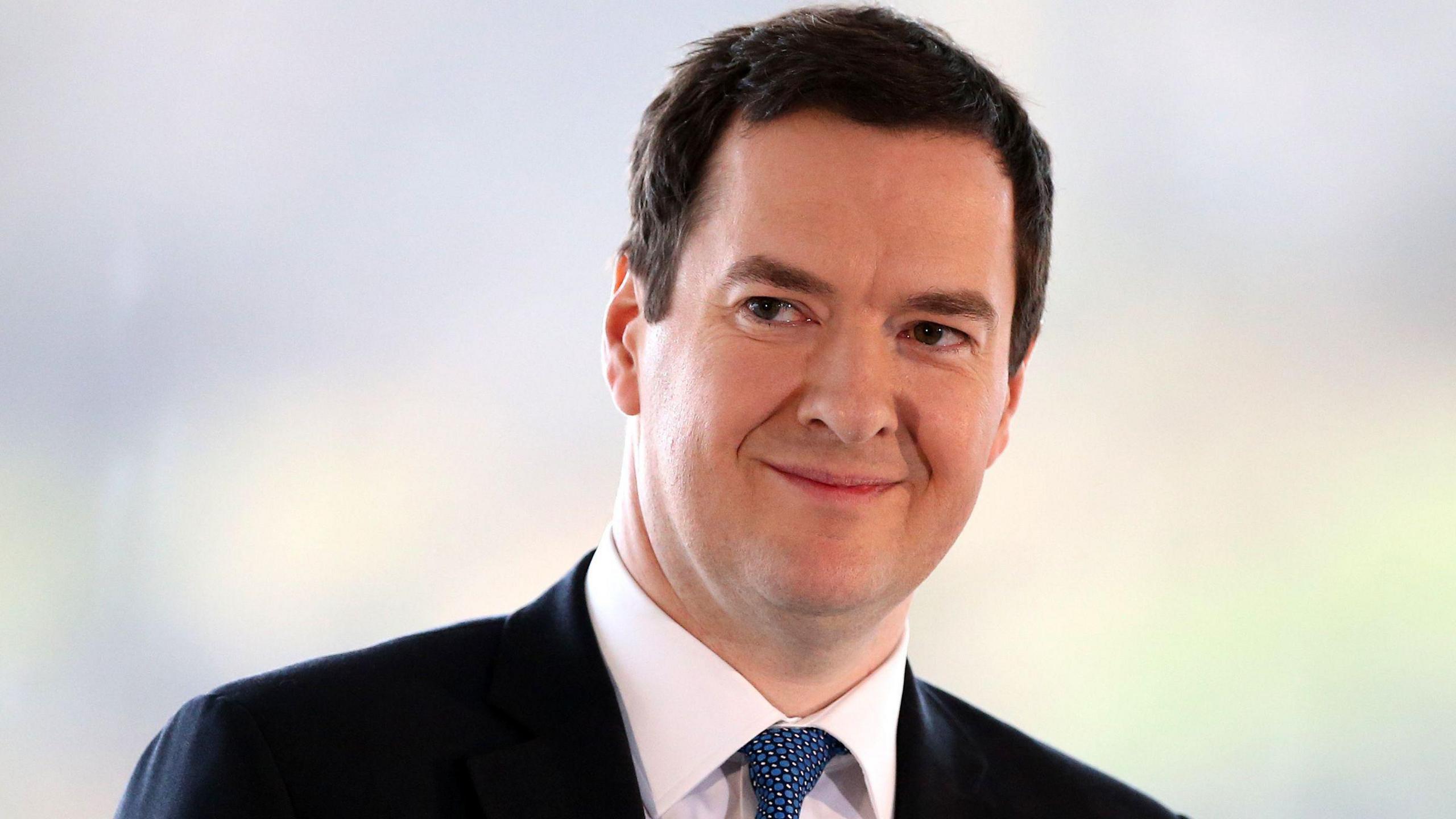 George Osborne, smiling, and looking off camera. He has short dark hair and is wearing a dark jacket, white shirt and blue patterned tie.