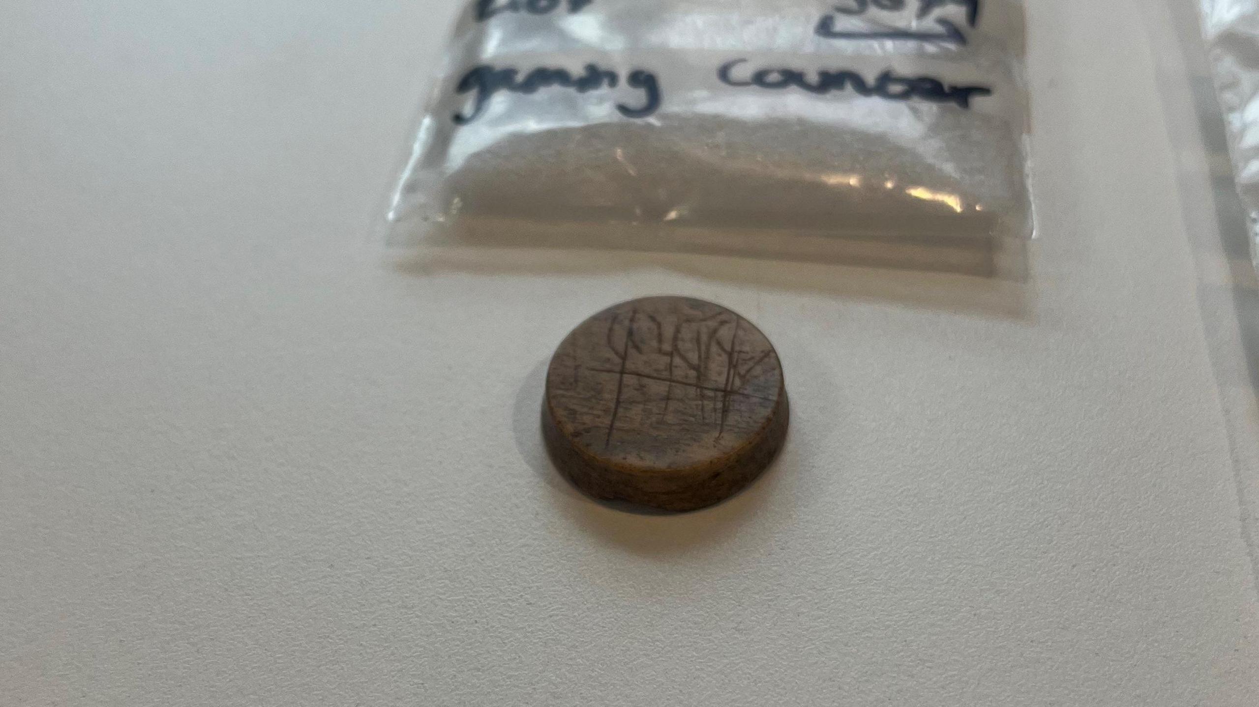 A round gaming counter. It is dark brown in colour and has some carvings on it. Above it is visible part of a plastic bag used to preserve it.