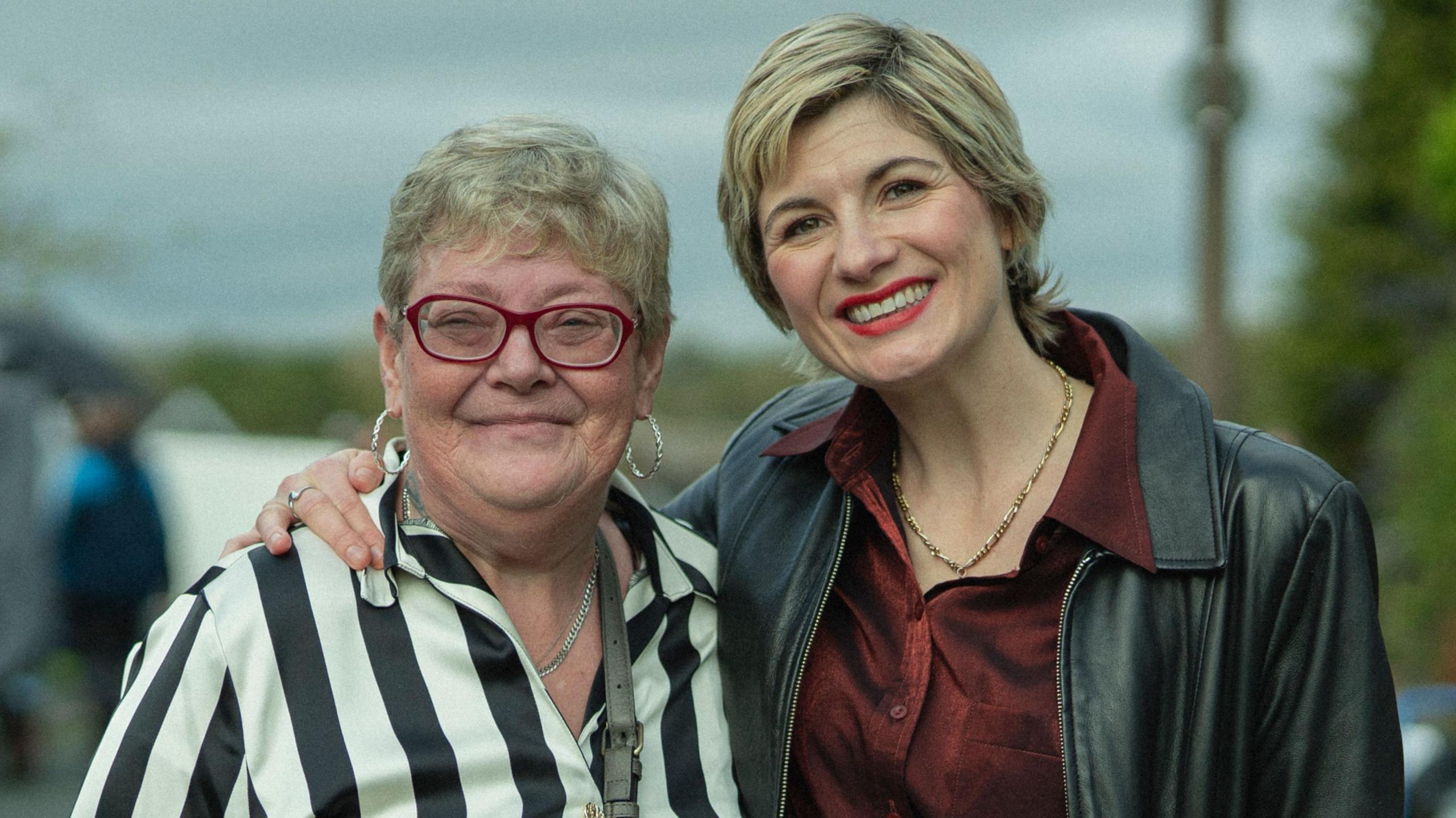 The real Susan McIntyre is standing with Jodie Whittaker. Both are smiling and Jodie has an arm around Susan.