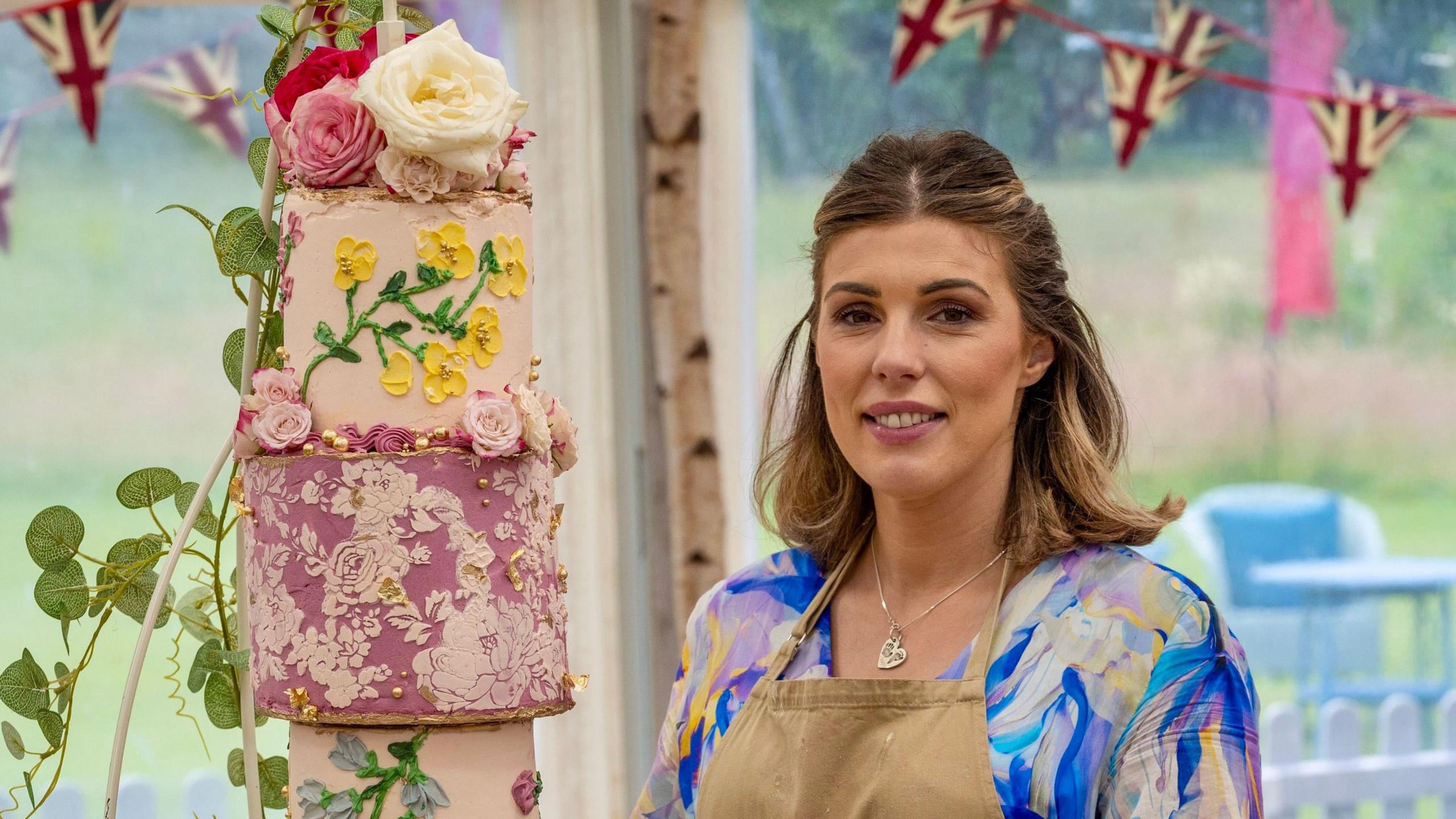 Georgie Grasso stood in the Bake Off tent. She is wearing a beige apron and underneath it has a blue, pink and white marbled top. She is looking at the camera with her brown and blonde hair half tied up and half down. There is a cake to the left of her, which is a three tiered cake, in different shades of pink with flowers adorned on it. 