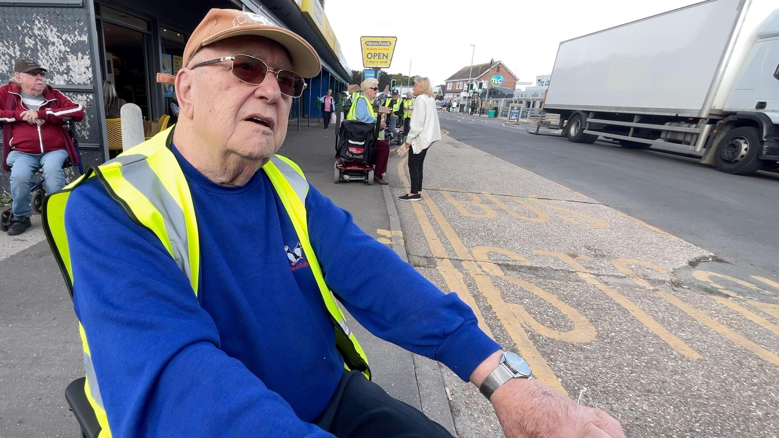 Len is wearing a blue jumper, a bright yellow Hi-Vis jacket and light orange cap with sunglasses on and is sat in a mobility scooter at the side of the road looking at the camera. There are more campaigners behind him at the side of the bus stop.