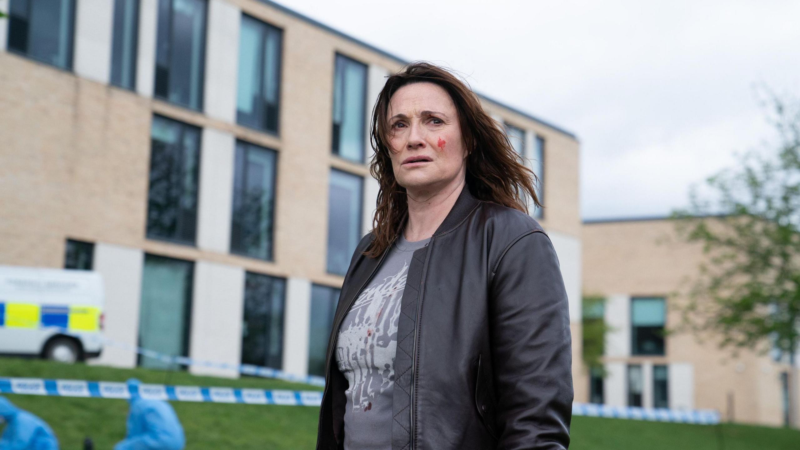 Sarah Parish stands outside a tall building, there is police tape, a police van and team in the shot. She has blood on her cheek and stares into the distance, looking worried.