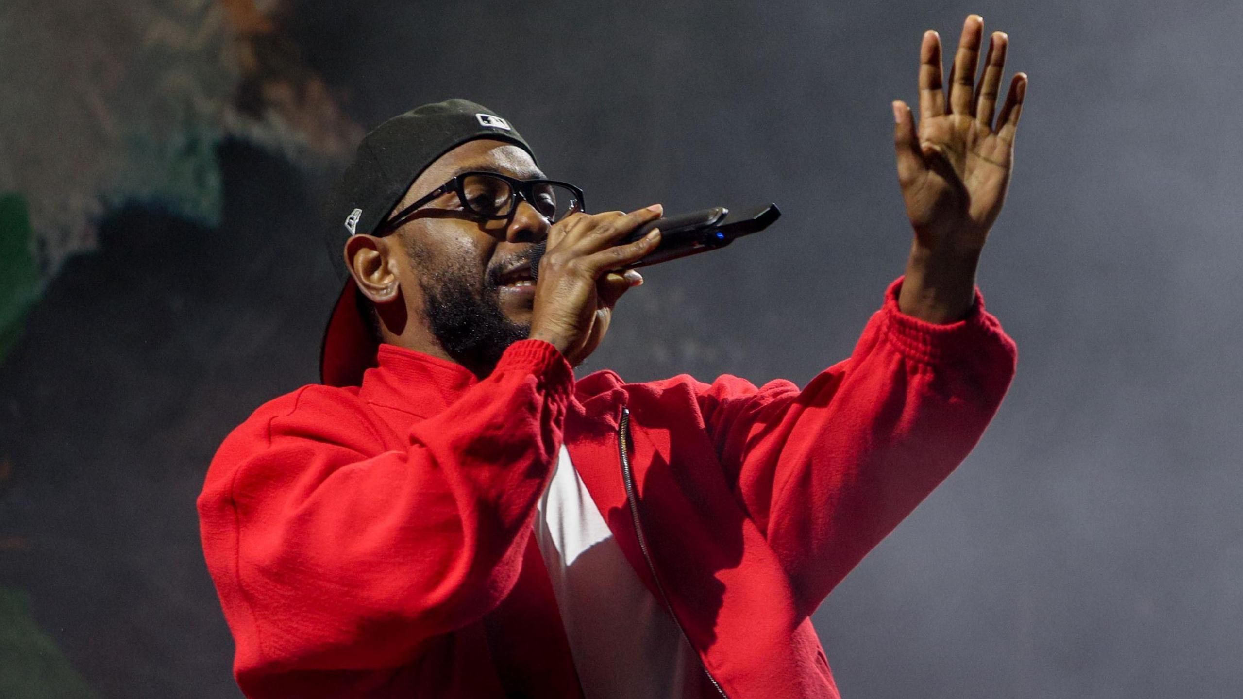 Kendrick Lamar performing, singing into a mic while holding his left hand up towards the audience. He wears a red jacket, black-rimmed glasses and a black baseball cap twisted backwards. 