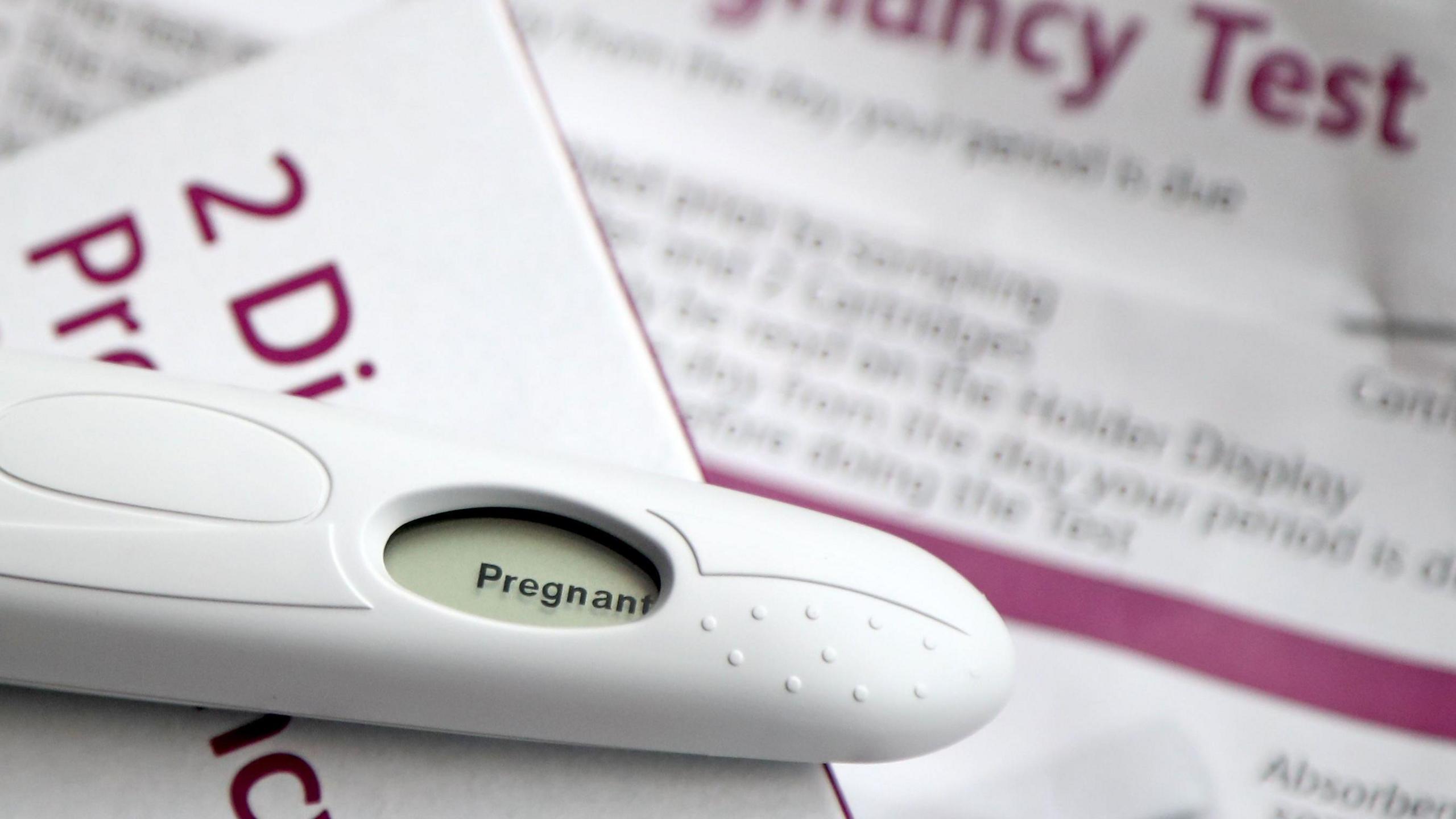 A positive pregnancy test lies on top of the test box which is white with magenta writing, the instructions for the kit are blurred out in the background.
