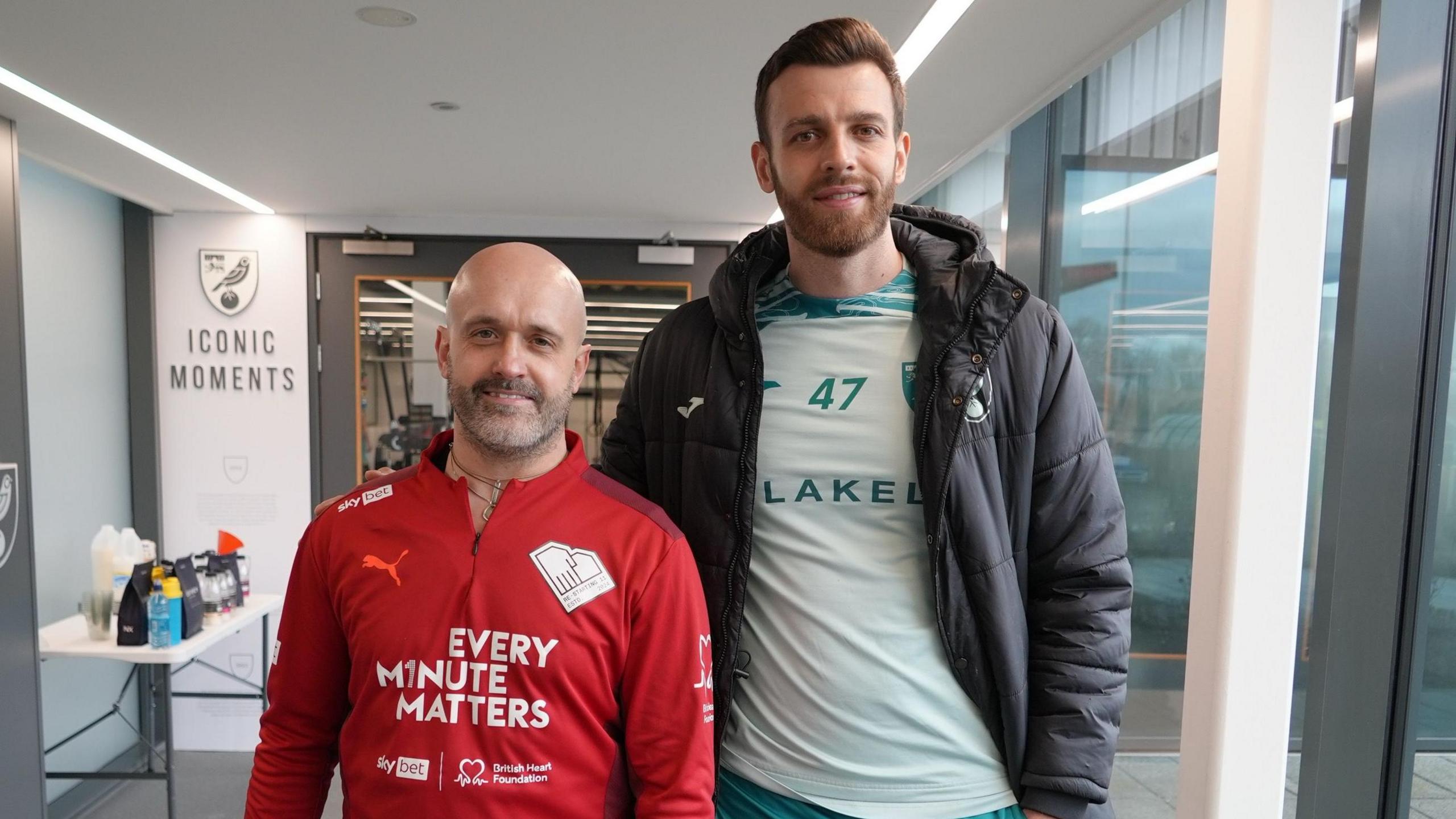 Andy Wall is standing next to Angus Gunn. They are in a brightly lit corridor at the Carrow Road football stadium. Mr Wall is wearing a red shirt which says EVERY MINUTE MATTERS, the slogan of a heart safety campaign. 