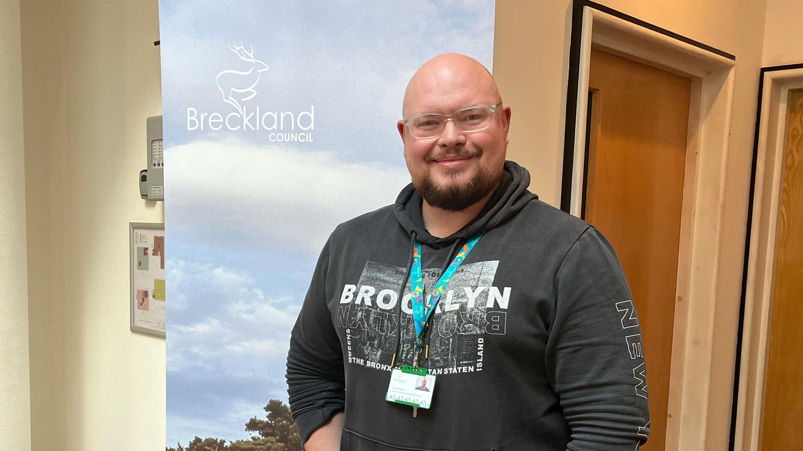 James Harding from Breckland Council stands in front of a banner which says "Breckland Council". He is bald with a beard, wearing a dark grey hoodie and jeans. 