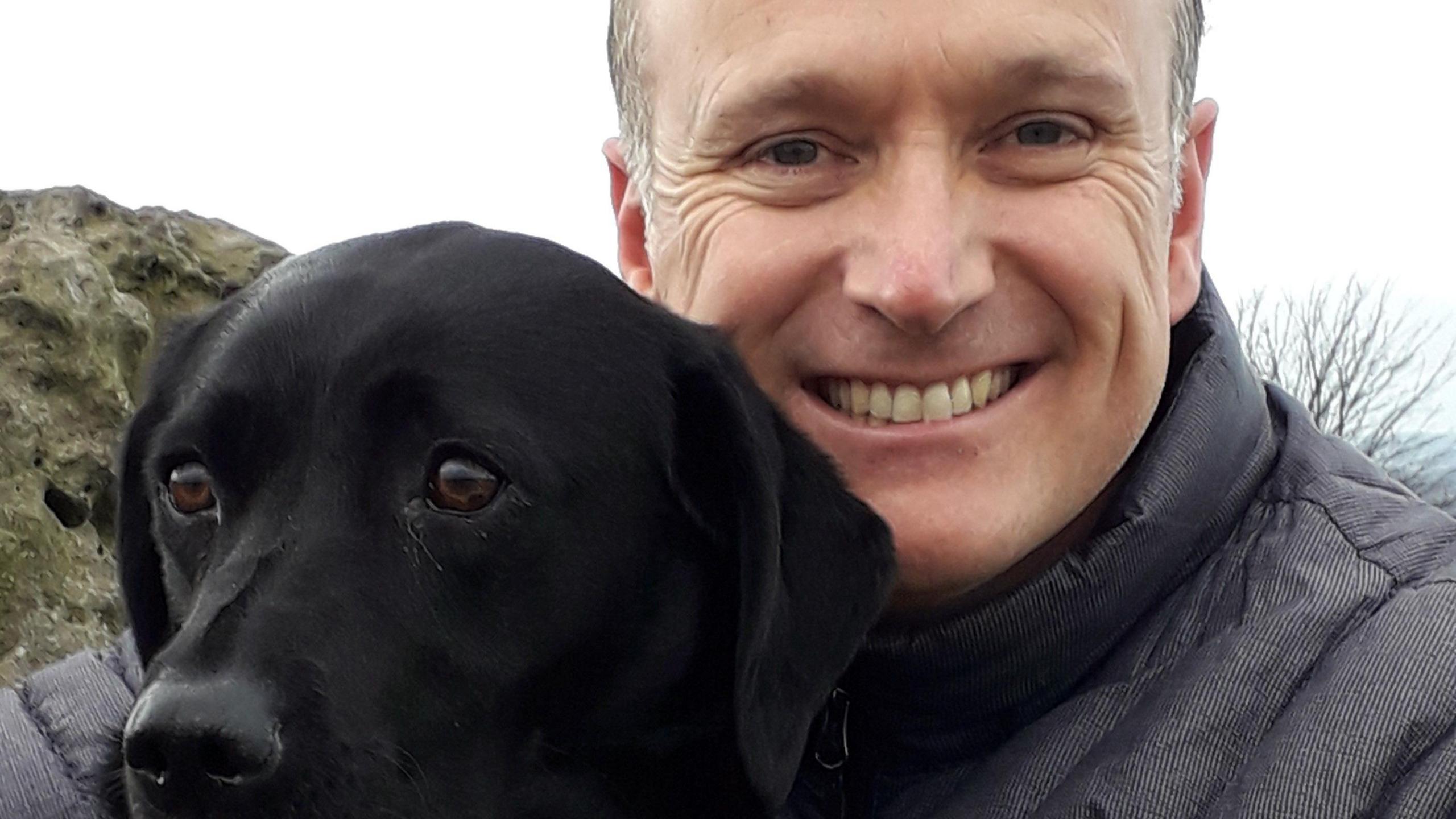 A man grinning while hugging a black dog, who is looking elsewhere