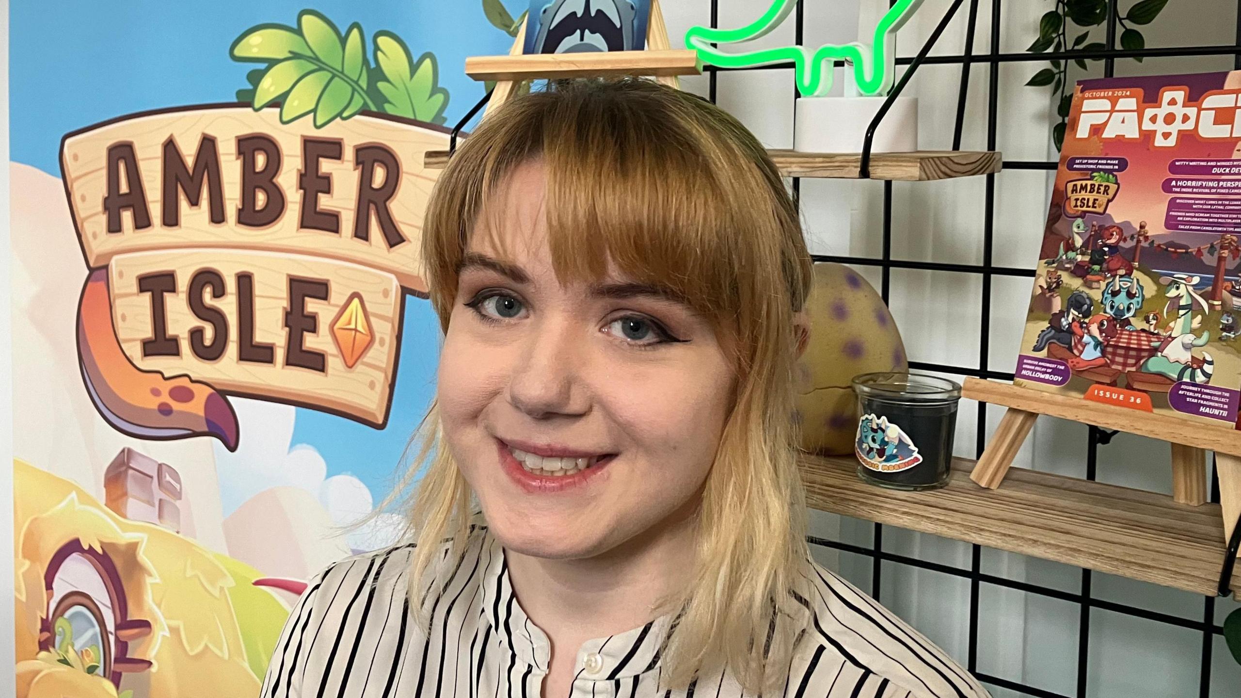 A young woman with blonde hair wearing a striped cream shirt. She is smiling at the camera and is stood in front of a banner which says 'Amber Isle' 