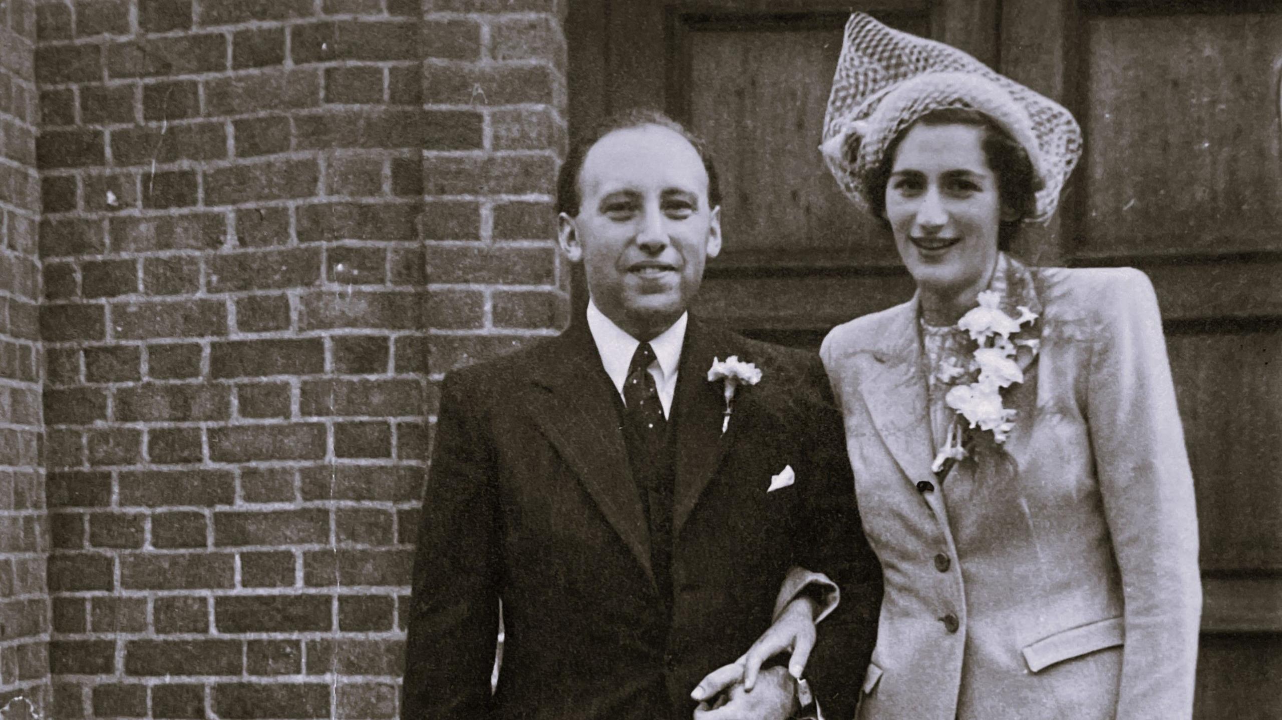 Llun du‑a‑gwyn o Alfred a Doris Moritz yn sefyll ochr yn ochr y tu allan i adeilad brics. Maent wedi gwisgo mewn dillad priodas ffurfiol: un ohonynt mewn siwt ddu gyda blodeuyn bach yn y lapel, a’r llall mewn siaced ysgafn gyda blodau ar y frest a het steilus gyda rhwyll. Mae drws pren mawr yn y cefndir, gan awgrymu eu bod yn sefyll y tu allan i leoliad seremoni neu adeilad swyddogol ar ddiwrnod arbennig.