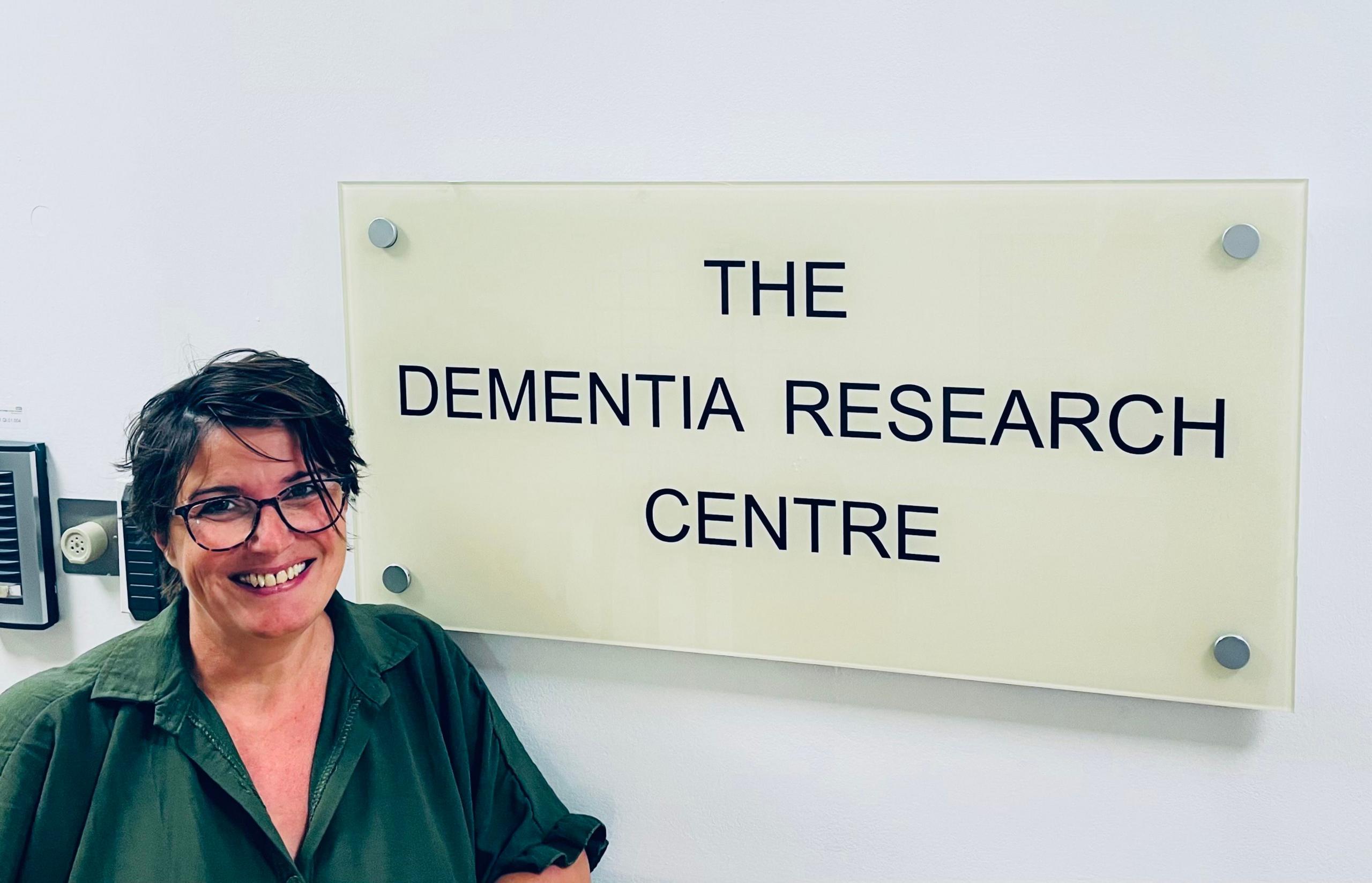 Sophie Leggett wearing glasses and a green shirt and is stood in front of a sign which reads "the dementia centre"