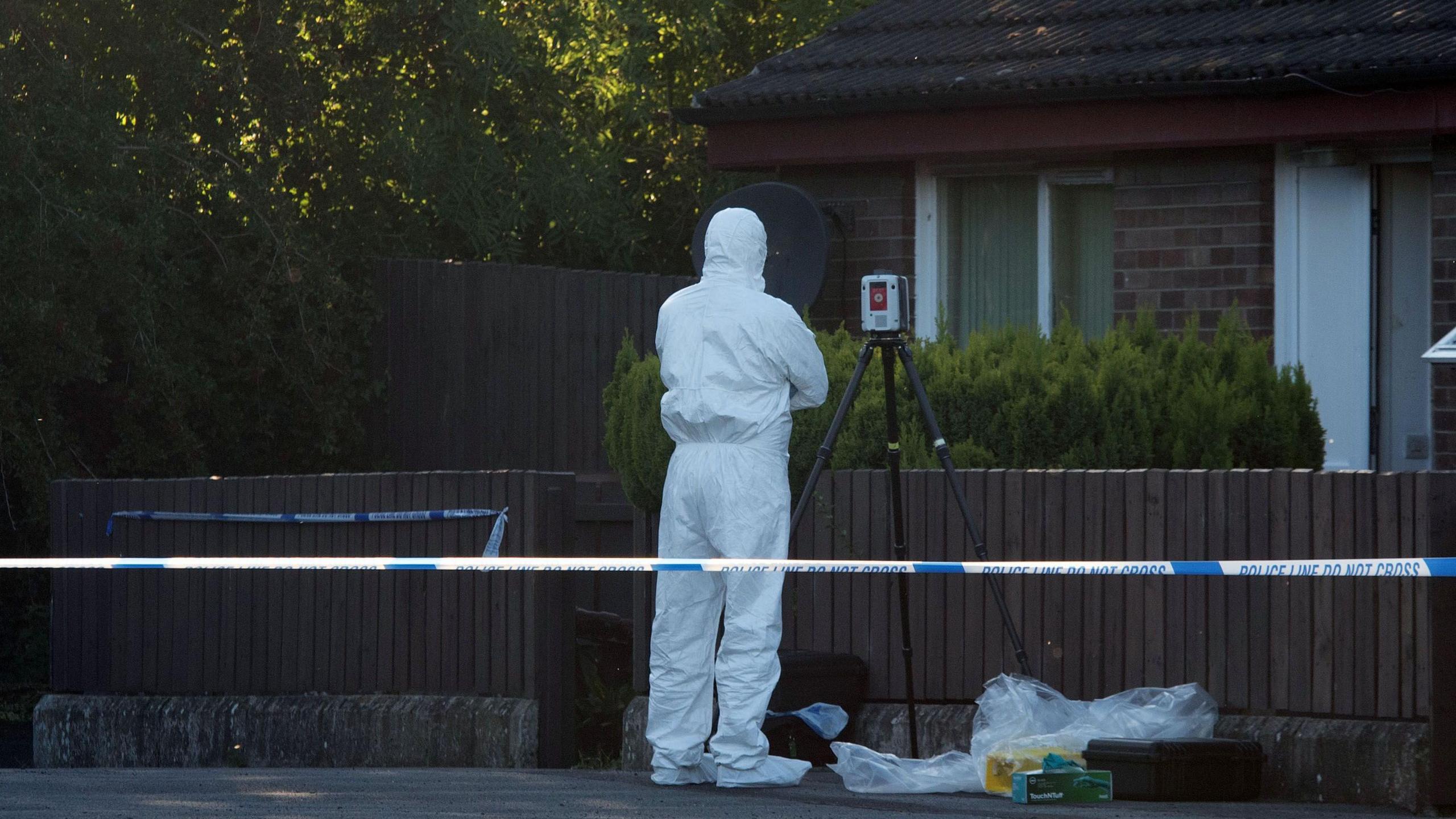 A man in a white forensic suit with a camera stands in front of a brown fence outside a red brick house. There are plastic bags on the ground beside him and a police cordon.