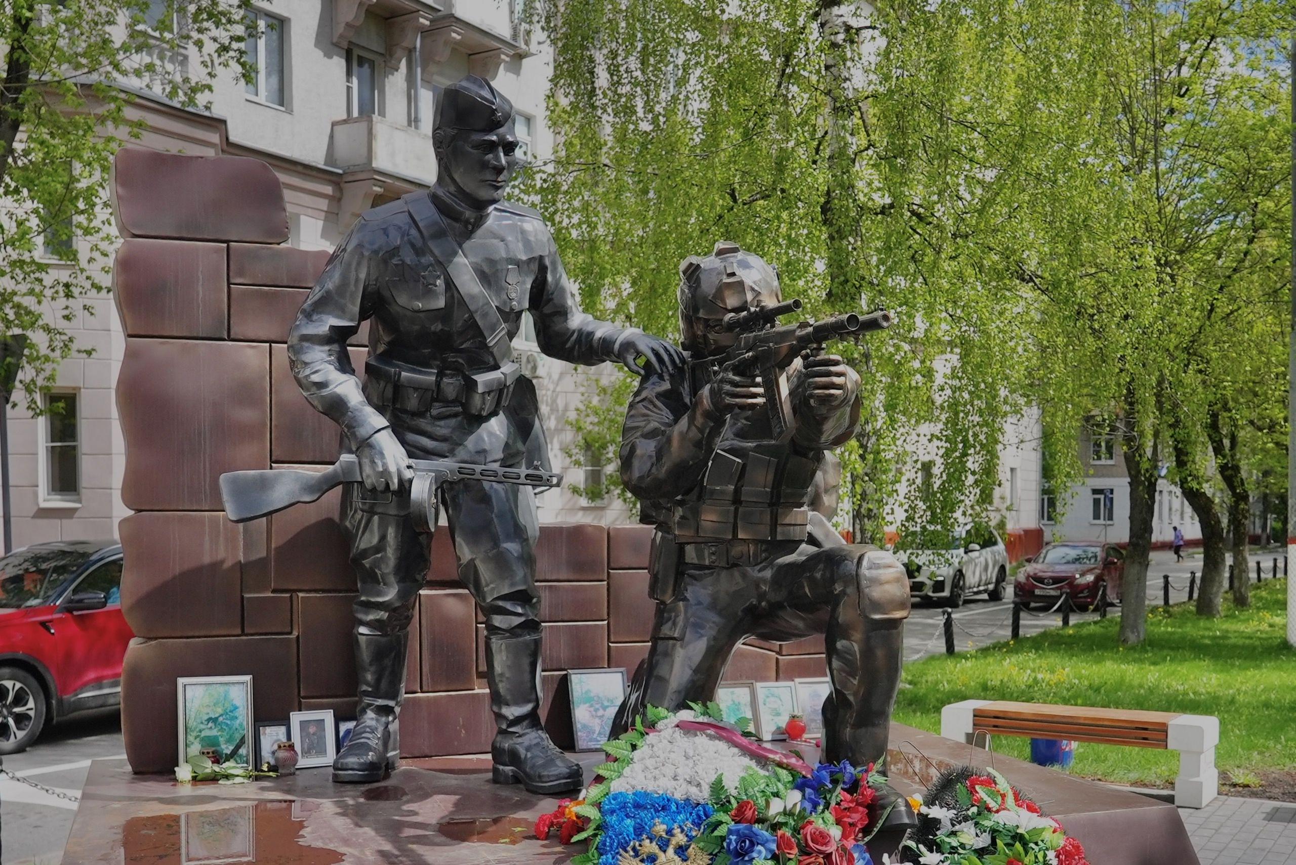 Two statues - one is a man wearing a military uniform from World War Two. He has his hand on the shoulder of a man wearing a modern military uniform, camos with a helmet, who is crouching and aiming a gun.