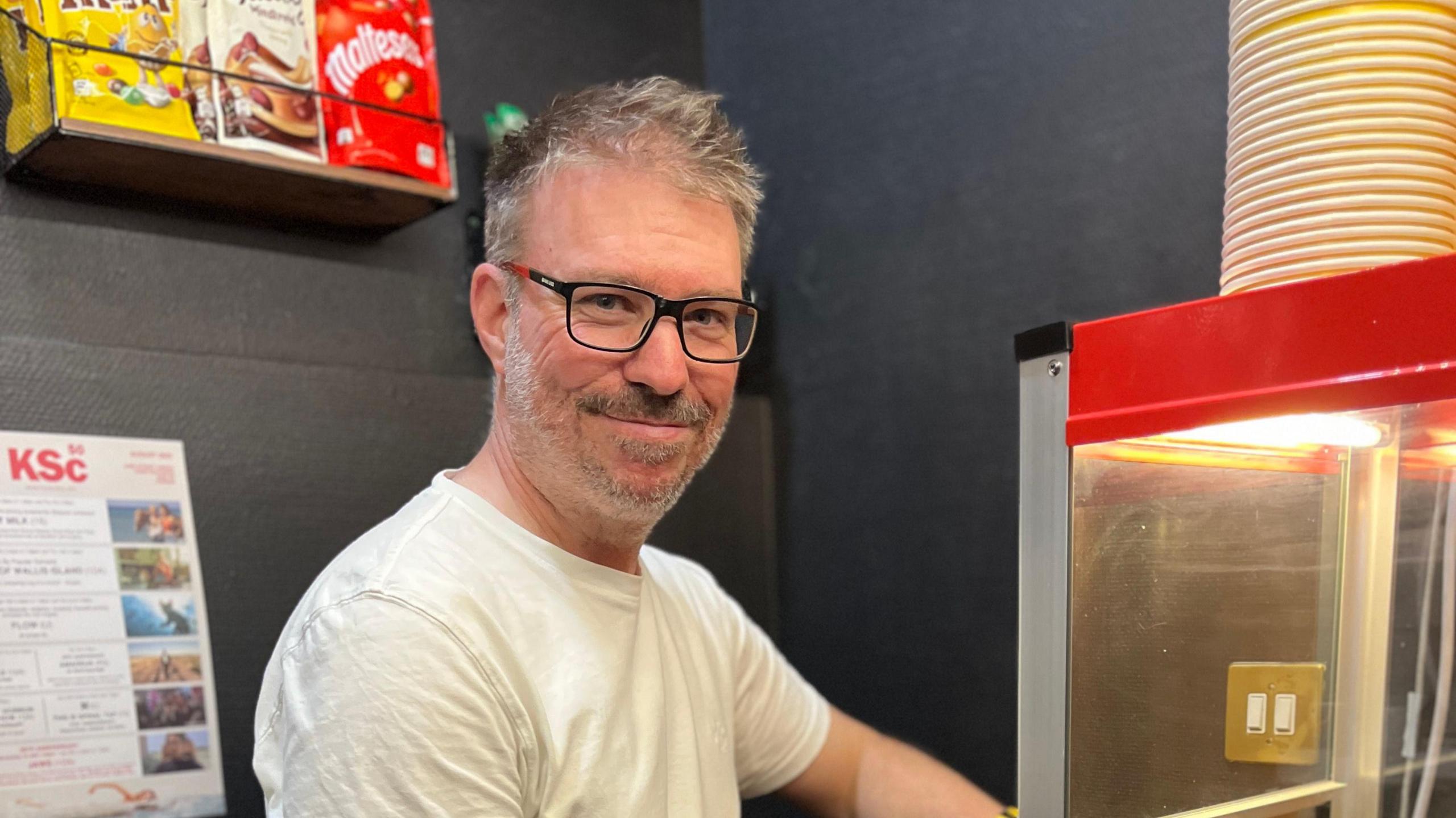 Mr Champion smiles at the camera next to a popcorn making machine. He has short dark hair with some grey in it and he wears a white T-shirt.