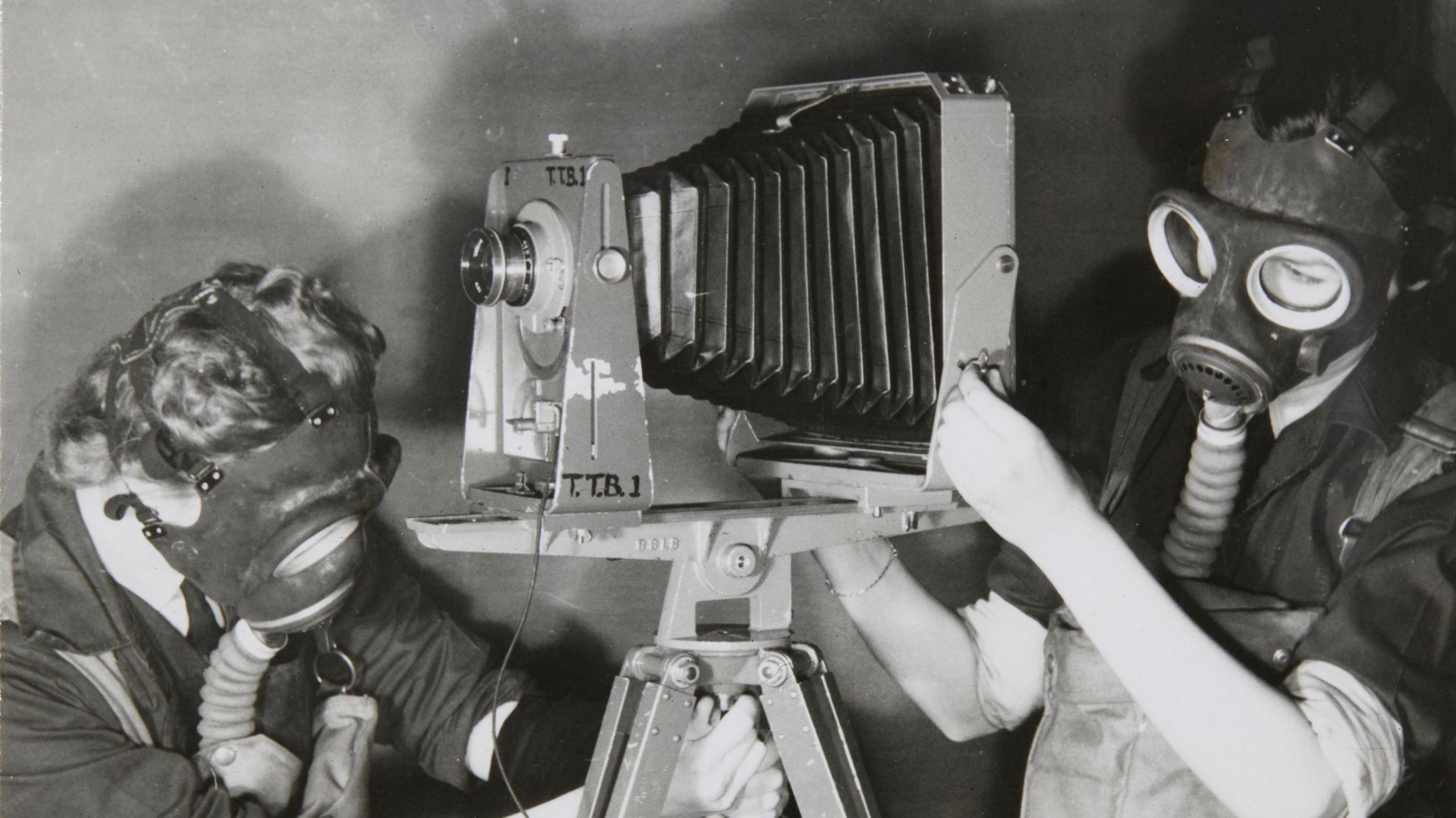 Two female trainees practising photography whilst wearing gas masks to simulate working under active service conditions. There are wearing unfiformed jackets and overalls. The photo is black and white.