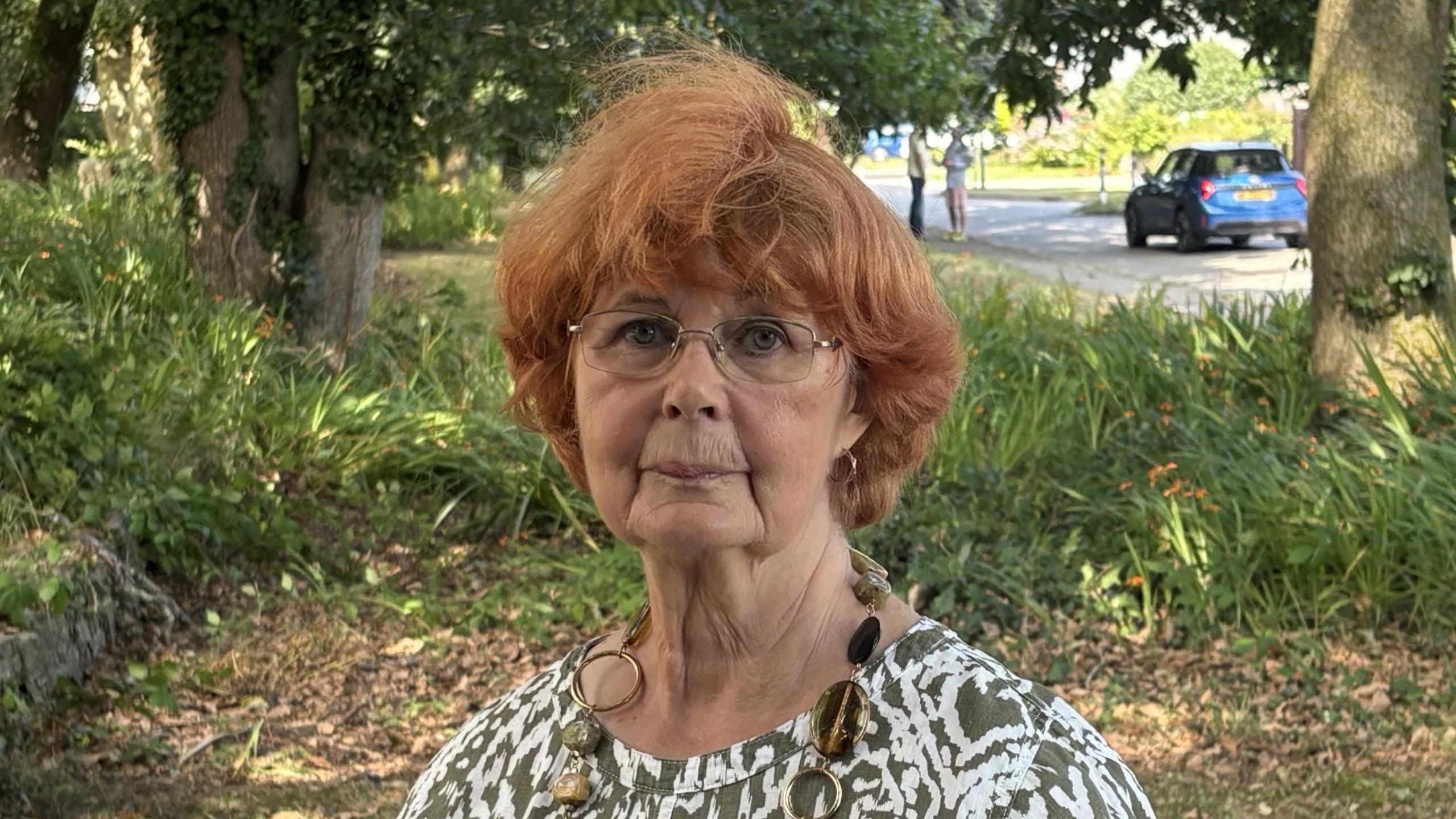 A woman with red hair and glasses wearing a green and white pattern top standing in a grassy area with trees in the background