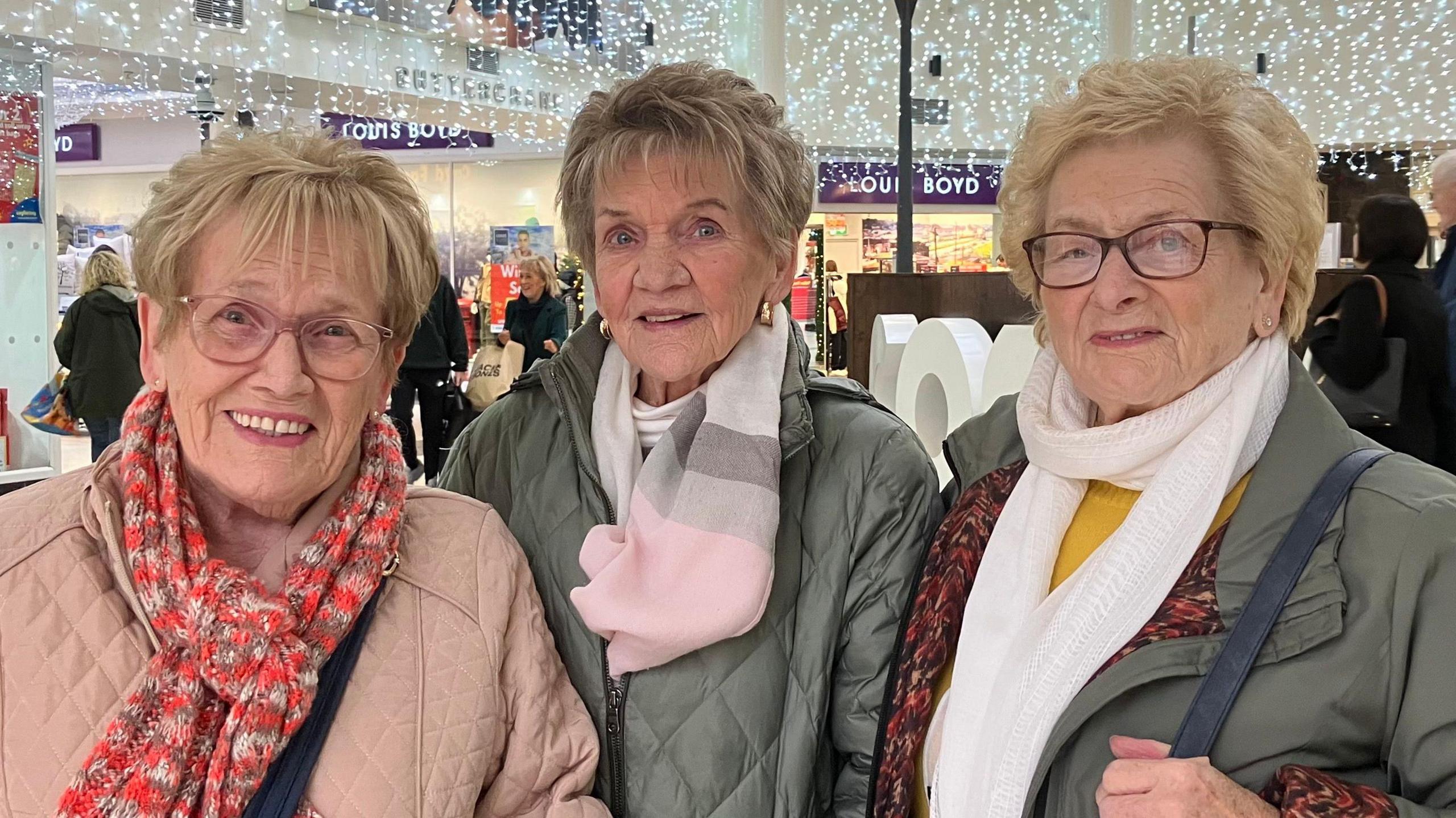 Three women are standing beside each other, each with a smile.  The woman on the left is wearing glasses, a pink coat and matching scarf.  The woman in the middle is wearing a grey padded jacket with a pink and grey scarf.  The third woman is also wearing a grey coat, yellow jumper and white scarf.  In the background there are other shoppers in this indoor shopping centre.  