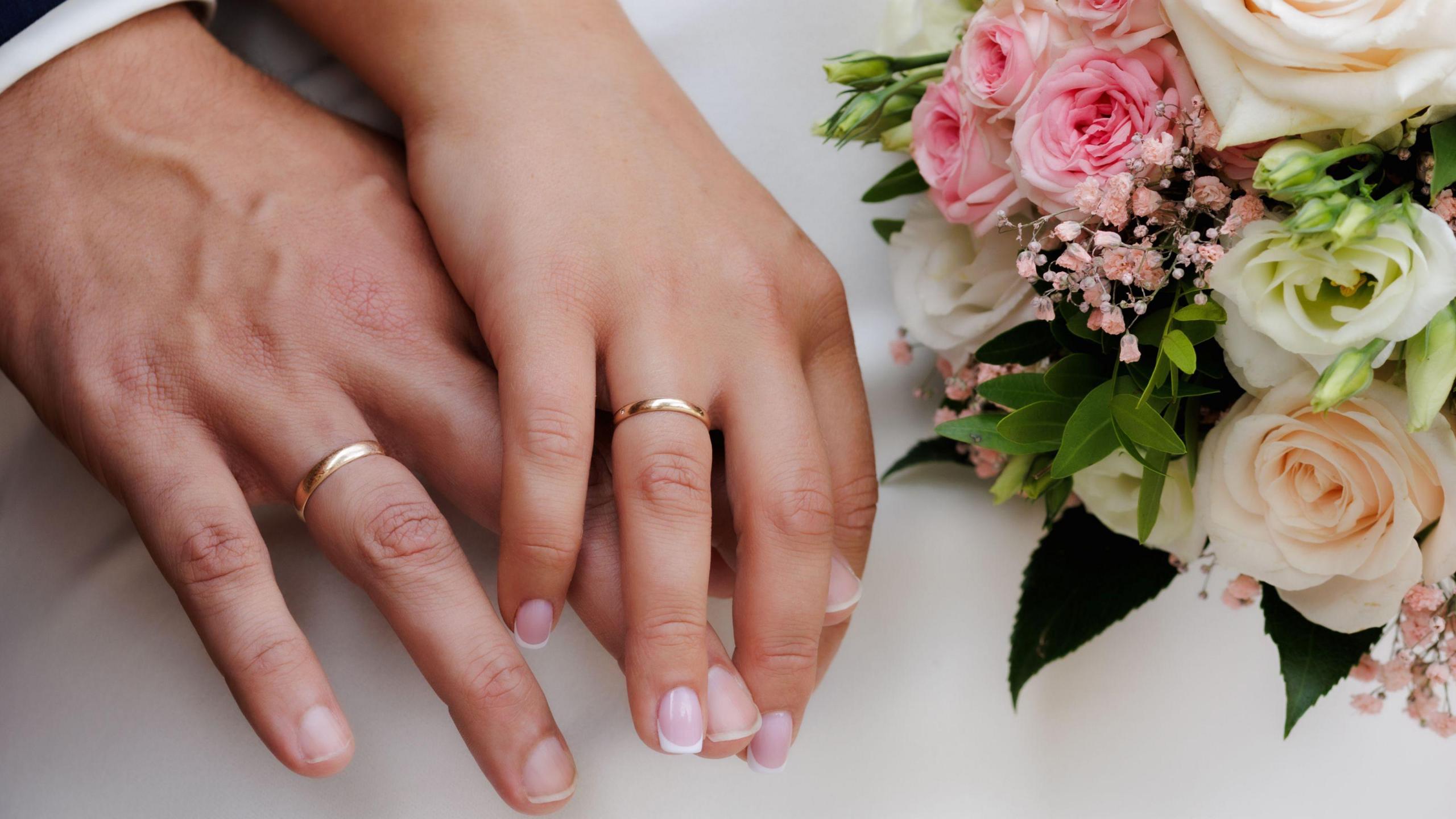 Two hands that have wedding rings on them. They are holding hands. Beside them is a bouquet of flowers