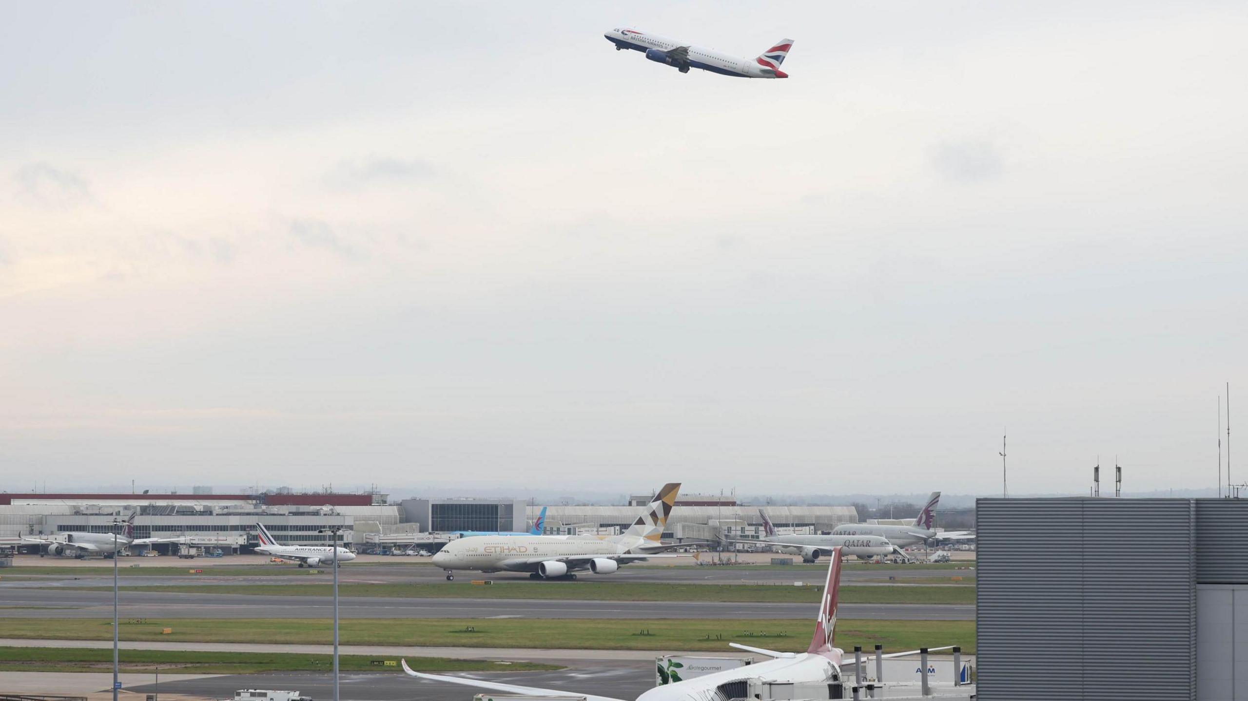 A view of Heathrow airport - several planes are sitting on runways, while one plane - a British Airways plane with a Union Jack design on the tail - is going into the sky.