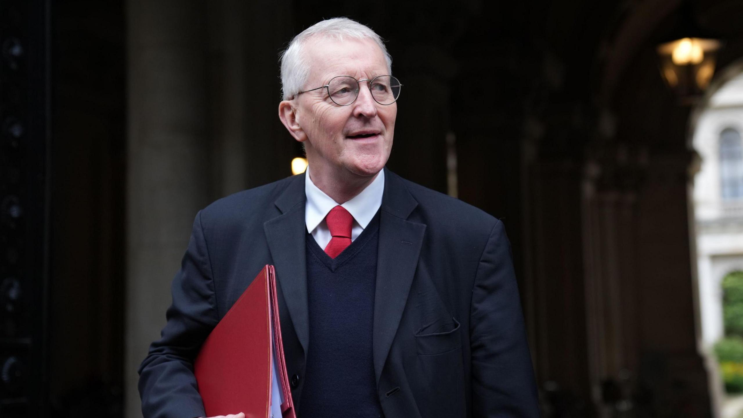 Hilary Benn - a man with white hair and circle glasses looks to the side as he walks through a stone archway with his mouth open. He is wearing a dark suit jacket, a v-neck jumper as well as a white collared shirt and red tie. He is carrying a red folder on his arm.