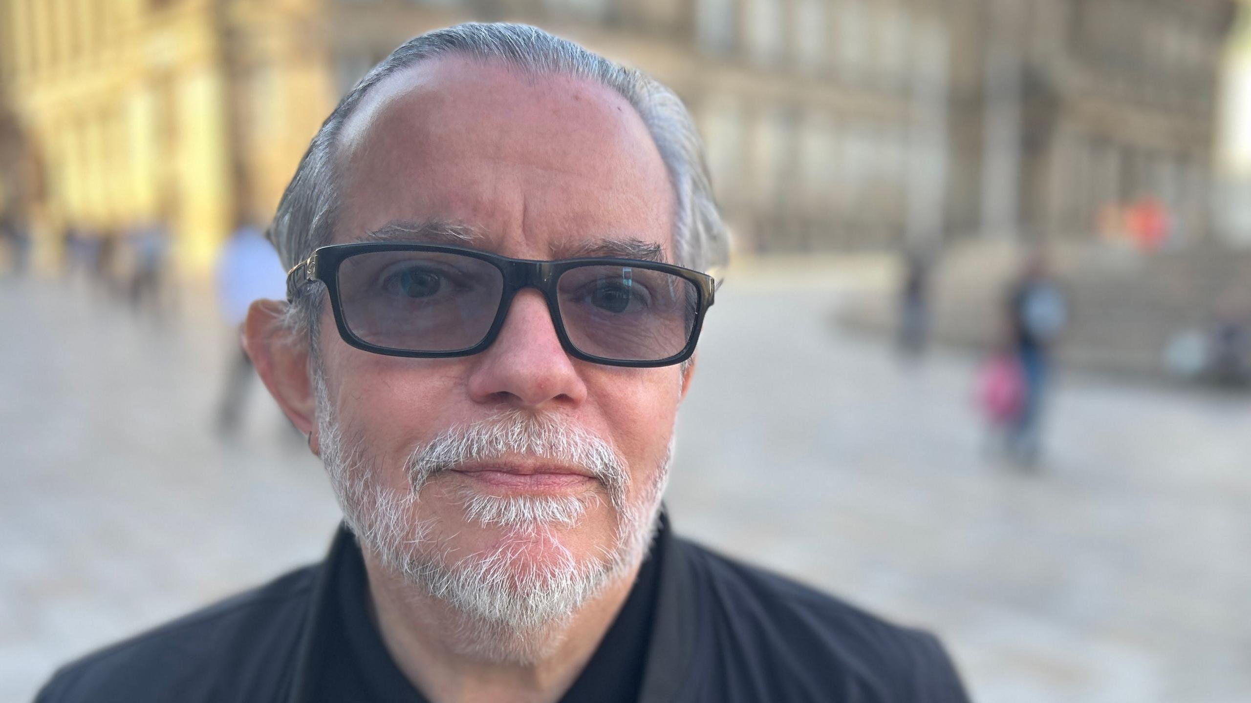A man with grey hair and grey beard wears black glasses and a black jacket with a black top underneath.