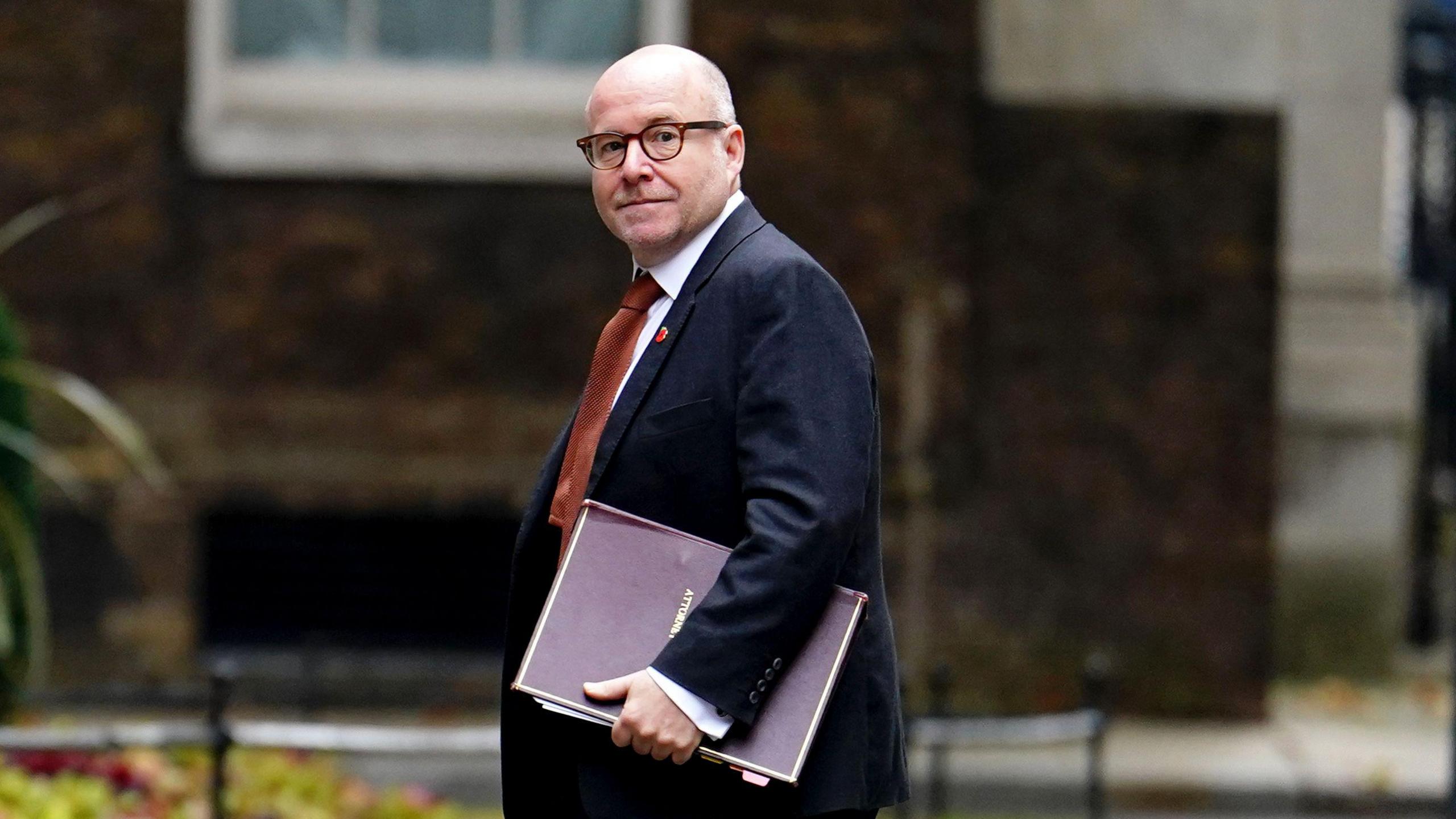 Attorney General Lord Richard Hermer arrives at Downing Street. He is wearing a dark suit and burnt orange tie with a small poppy on his left lapel. He is carrying a dark red file in his hand.