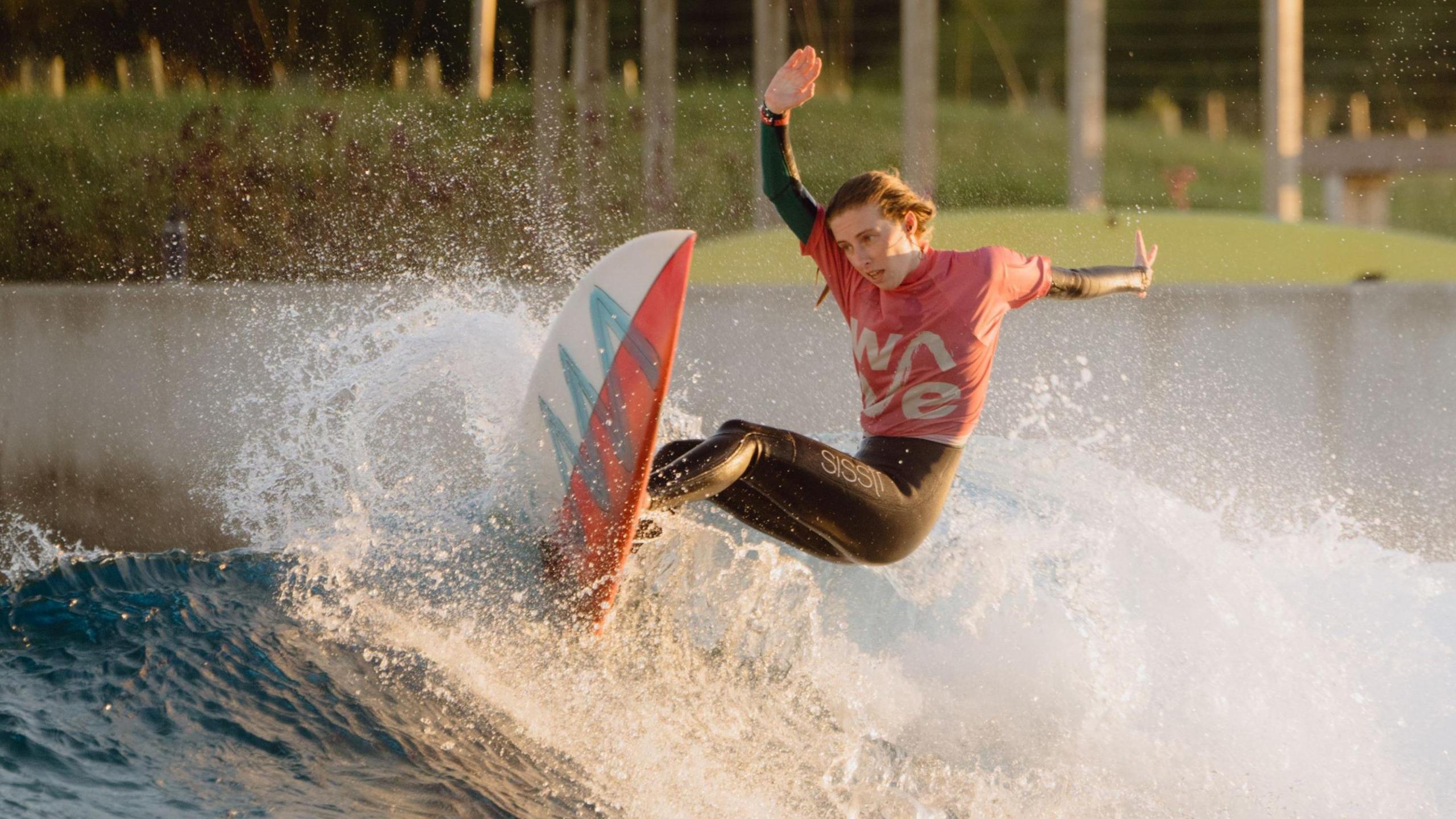 A young woman in a bright-coloured wetsuit and with long hair surfs the crest of a breaking wave at man-made surfing lake The Wave in Bristol. There is foaming white water at the crest of the wave.