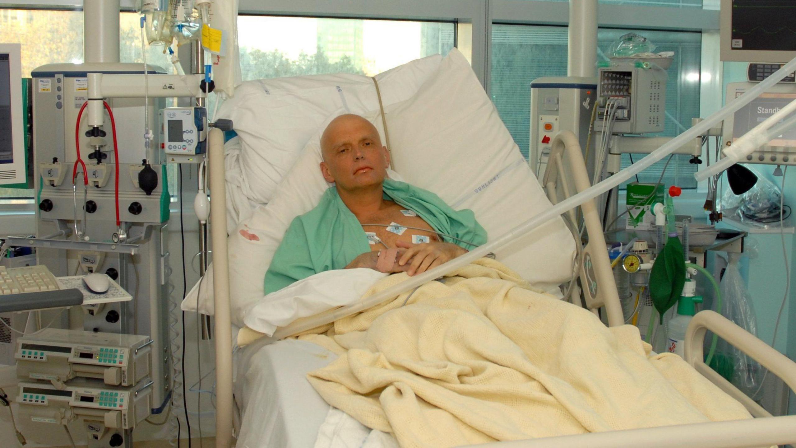 Alexander Litvinenko lying in a hospital bed at the Intensive Care Unit of University College Hospital in London on 20 November 2006. There are medical machines around the bed and he is wearing a green hospital gown. He is bald and tubes can be seen attached to his chest.