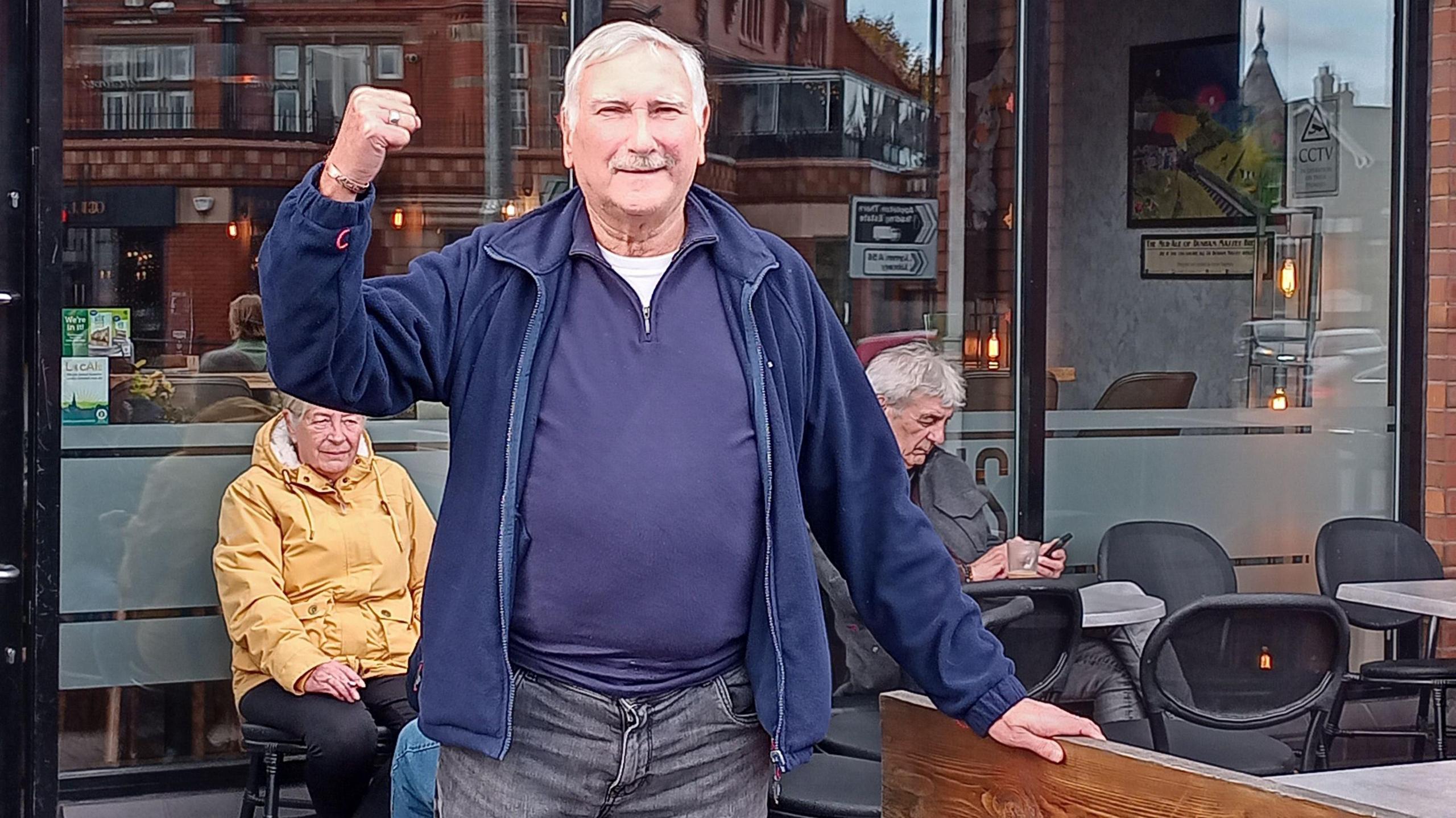 A man with short white hair and a moustache, wearing a blue jacket, jumper and jeans, raises his right fist in the air in celebration while standing outside a pub.