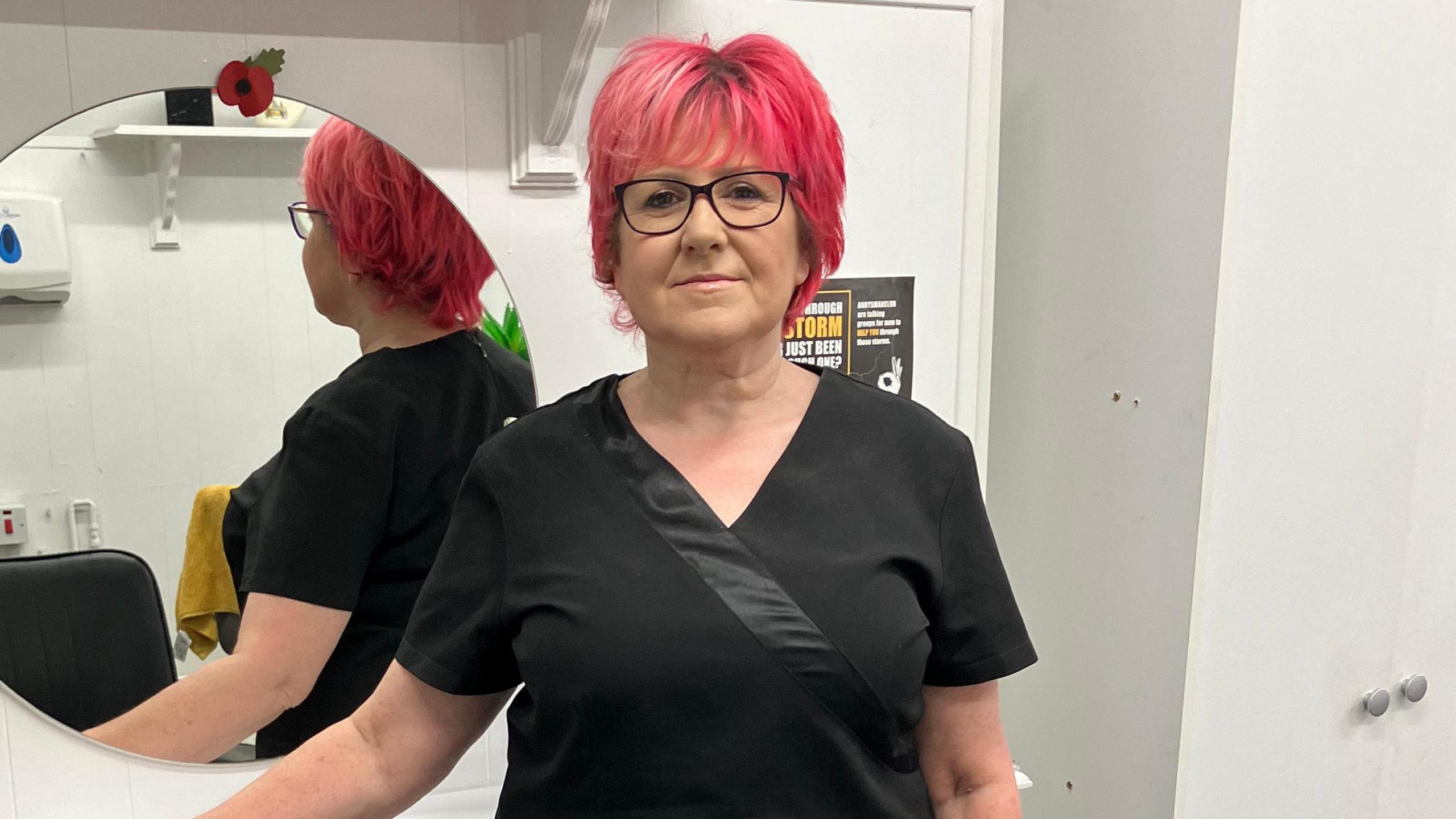 Petra Brown leaning against one of the tables in her barber shop. She has red hair, is wearing glasses, and a black top.