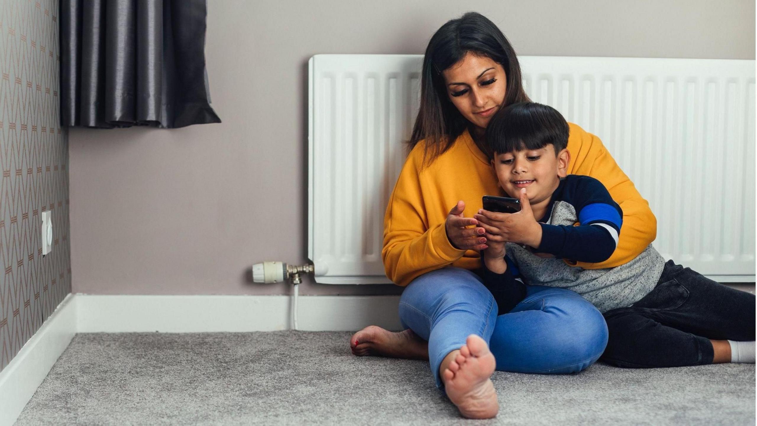 A mother and her child sit on the floor in front of a radiator. She has her arm around him and they are both looking at a phone. She is wearing a mustard-coloured top and blue denim jeans. The room in view is carpeted but otherwise bare. This is a stock picture, posed by models.