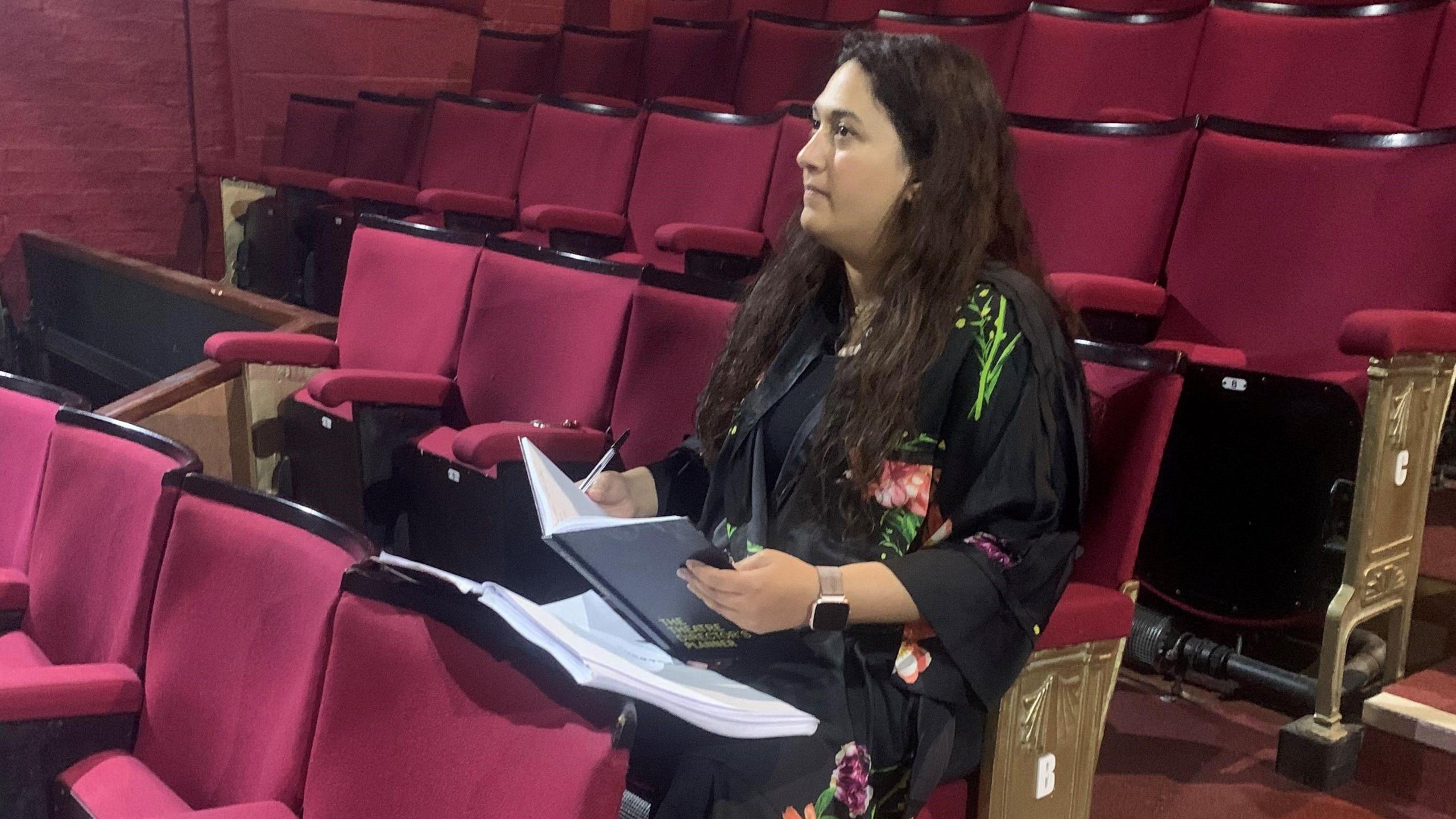 A woman with long dark hair sitting on a red theatre chair holding a notebook and pen as she watches the rehearsal