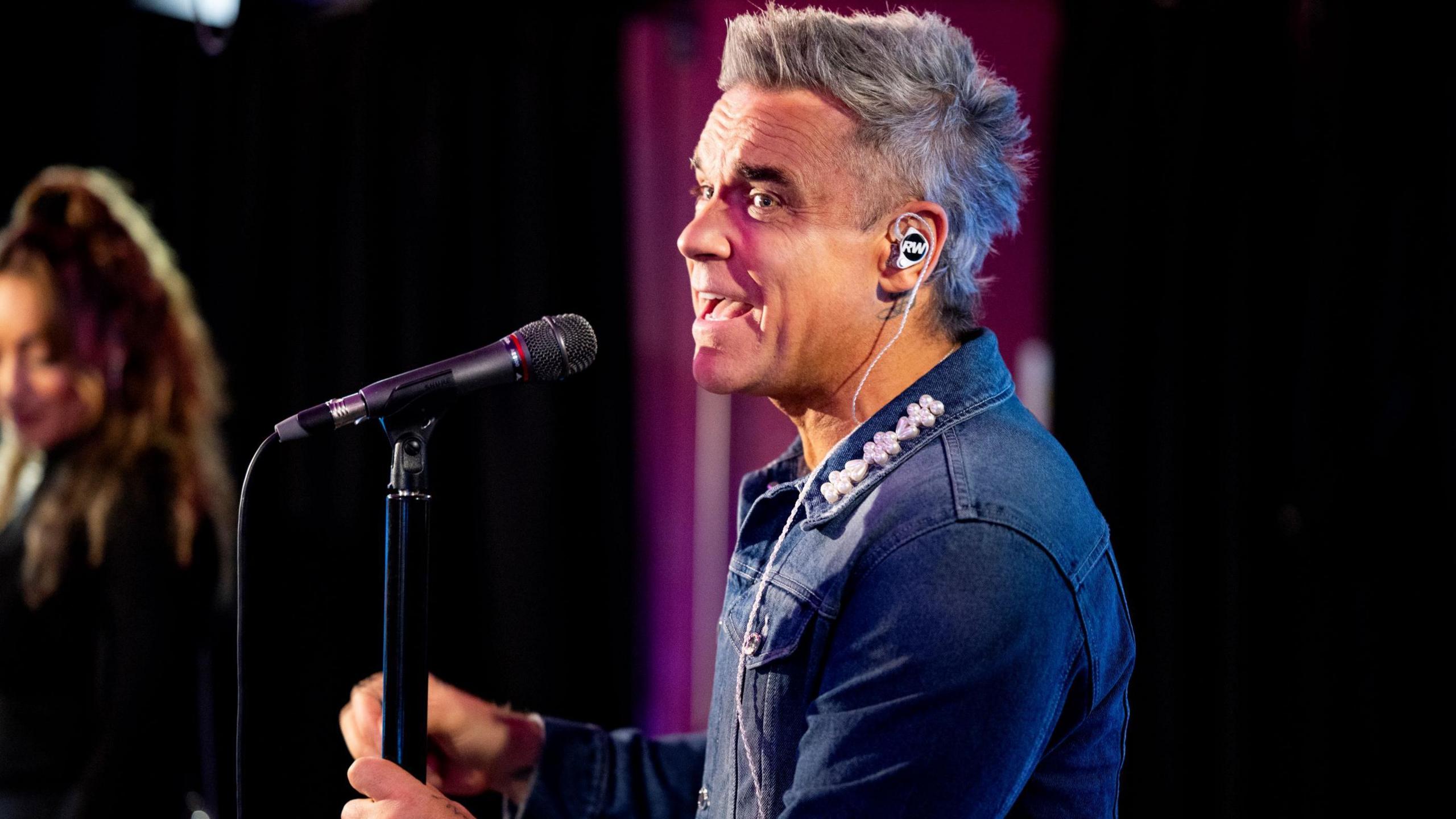 Robbie Williams singing into a microphone. He is wearing a jean shirt with a white collar and has grey spiky hair. 