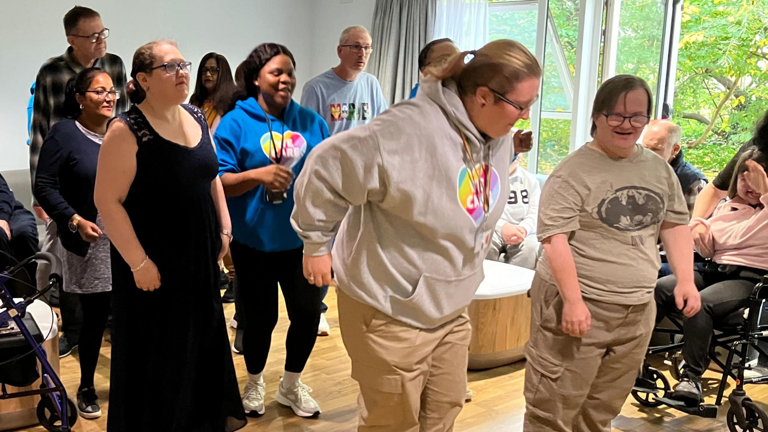 The picture shows a group of around eight people dancing in lines. A couple of people can be seen sitting in the background. They are in a room with a light brown wooden floor and a white wall behind them. To the side there are large patio doors.