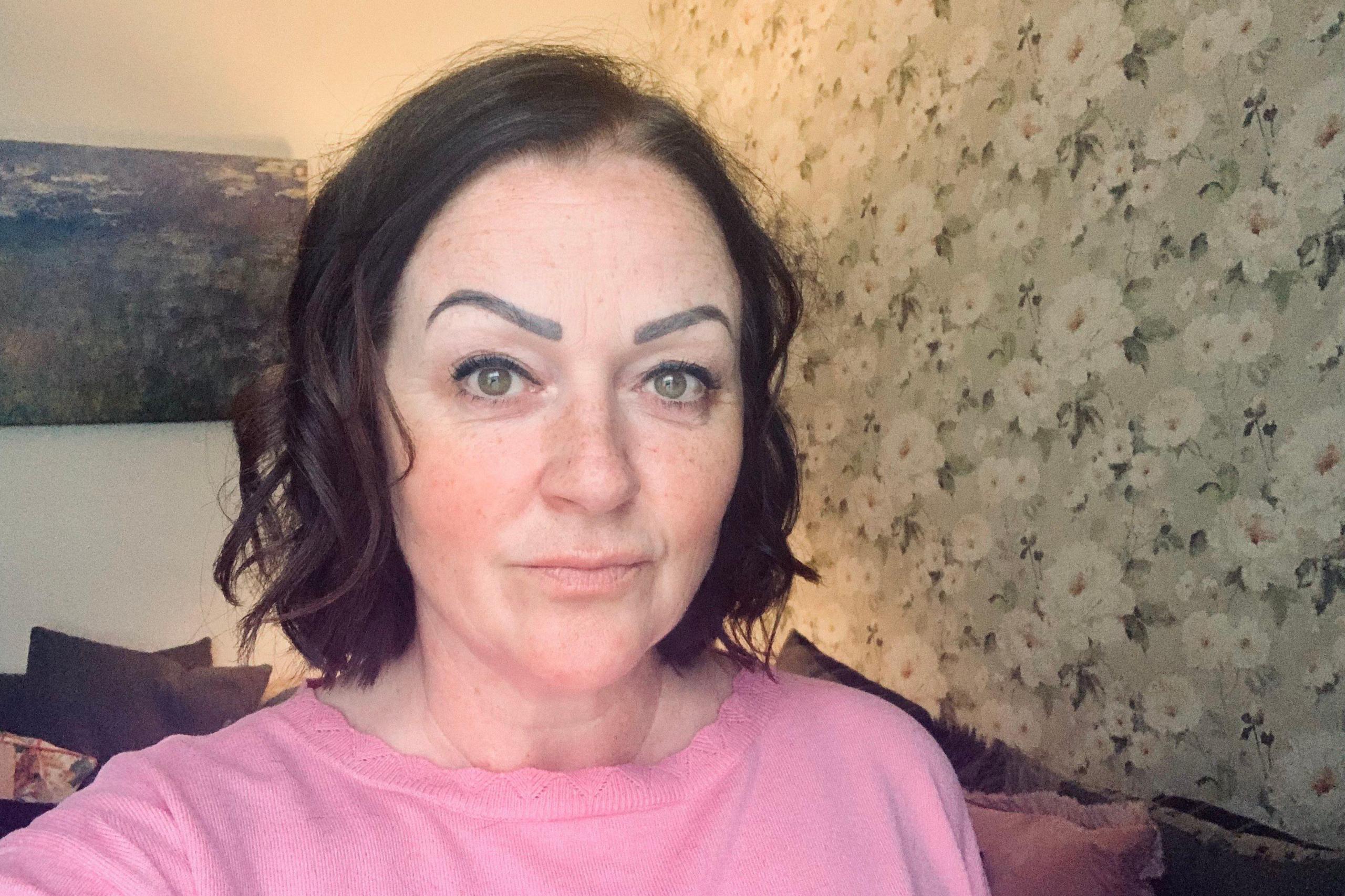 Woman standing in front of a wallpapered wall, wearing a pink top and looking into the camera