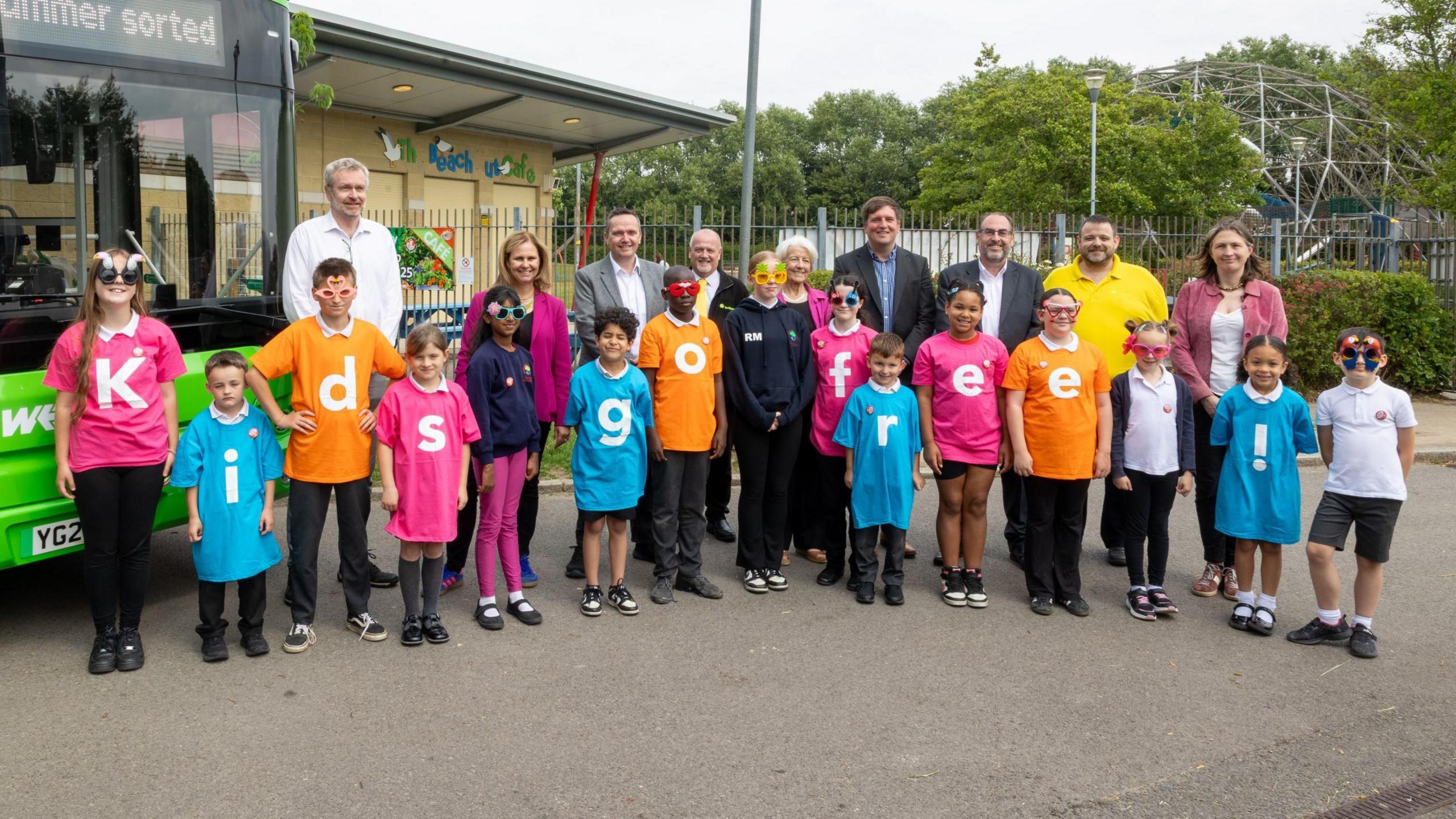 A group of children and adults pose in two lines in front of a green single-decker bus. The children's tops each have a letter on spelling out "Kids Go Free" as part of a summer bus promotion in Bristol