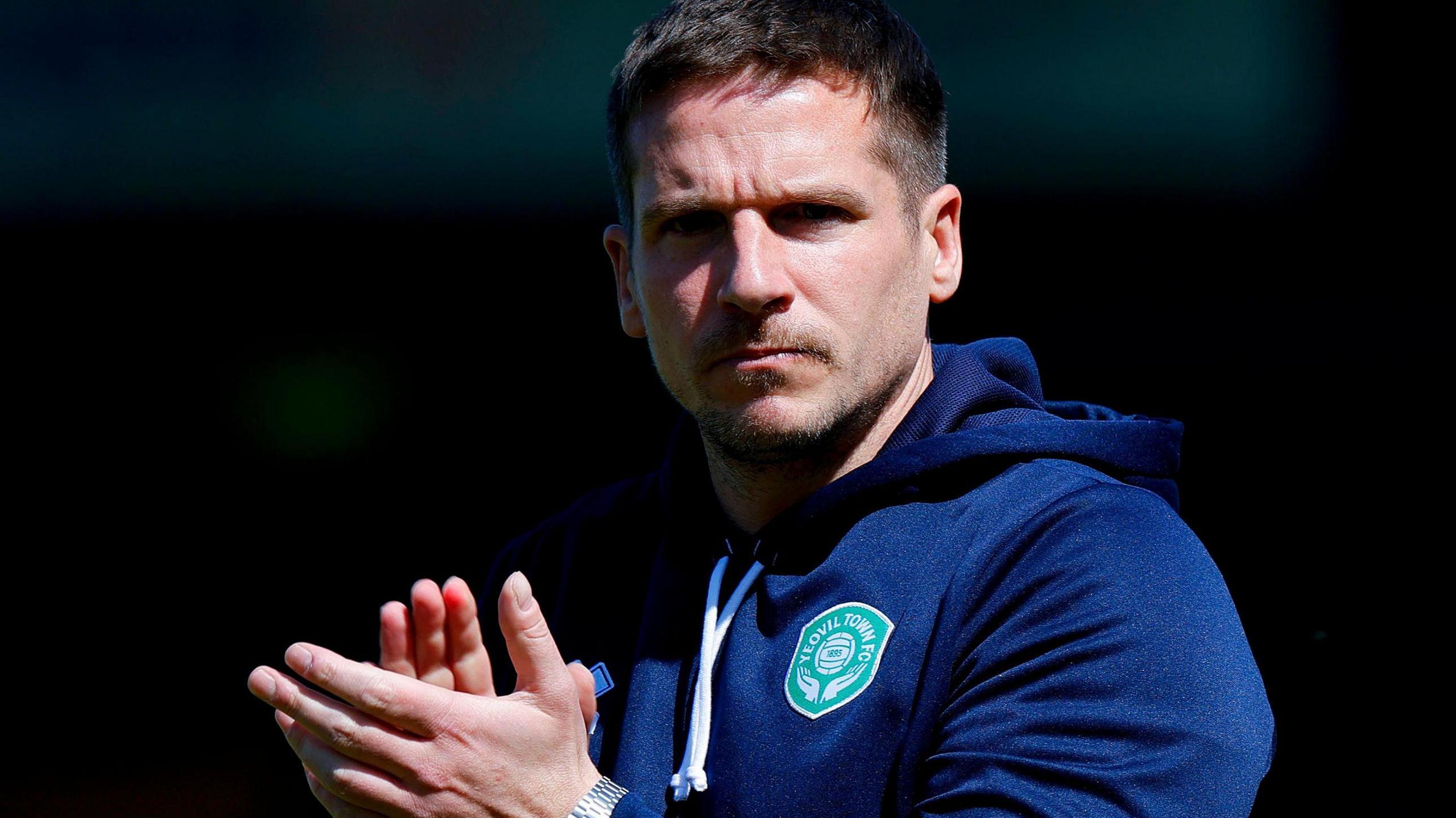 Yeovil Town manager Billy Rowley, wearing a navy blue hoodie with the club's crest on it, claps his hands